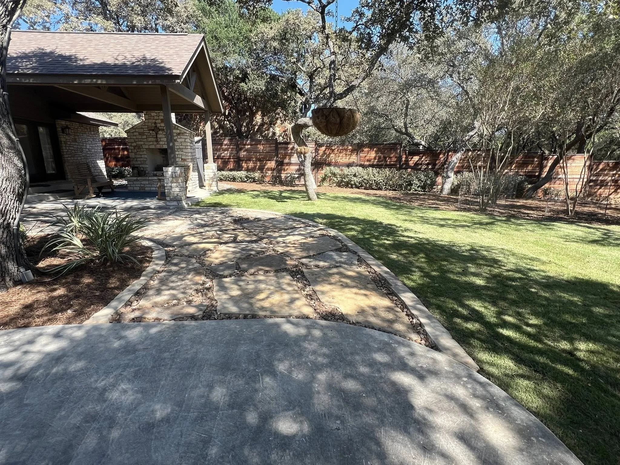 A backyard with a curved stone and concrete pathway, a grassy lawn, trees, and a brick house with a chimney and a porch.