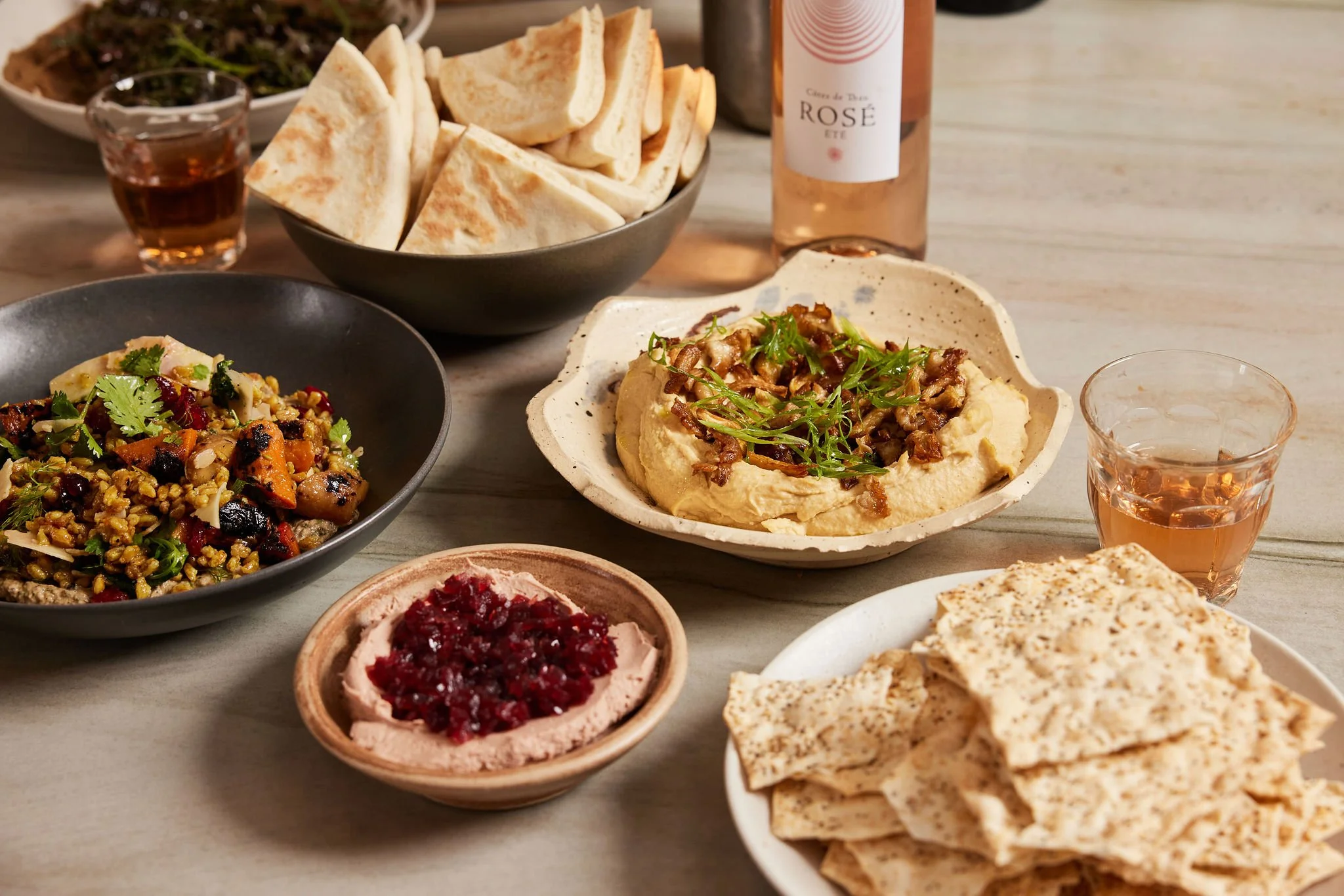 An assortment of Middle Eastern dishes on a wooden table, including hummus topped with chopped nuts and herbs, a bowl of tabbouleh, a bowl of pita bread, a small dish of rose wine, and glasses of pink wine.