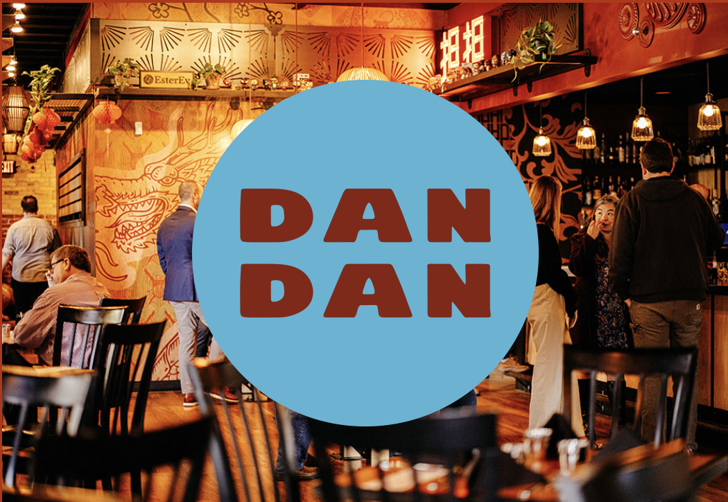 Interior of the DanDan restaurant with people socializing, wooden decor, Asian-inspired wall art, hanging lanterns, and a large blue circle with the text 'DAN DAN' in brown.