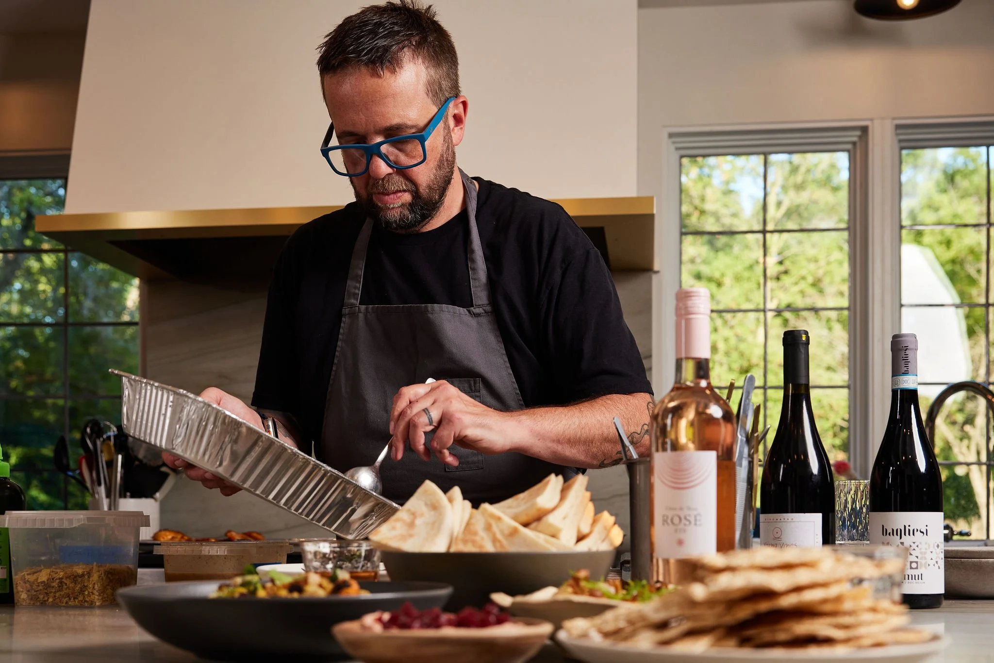 Chef Dan Jacobs preparing food in a bright kitchen. He is wearing a black T-shirt and a gray apron. The kitchen has large windows, and the counter is filled with plates of bread, wine bottles, and various food items.