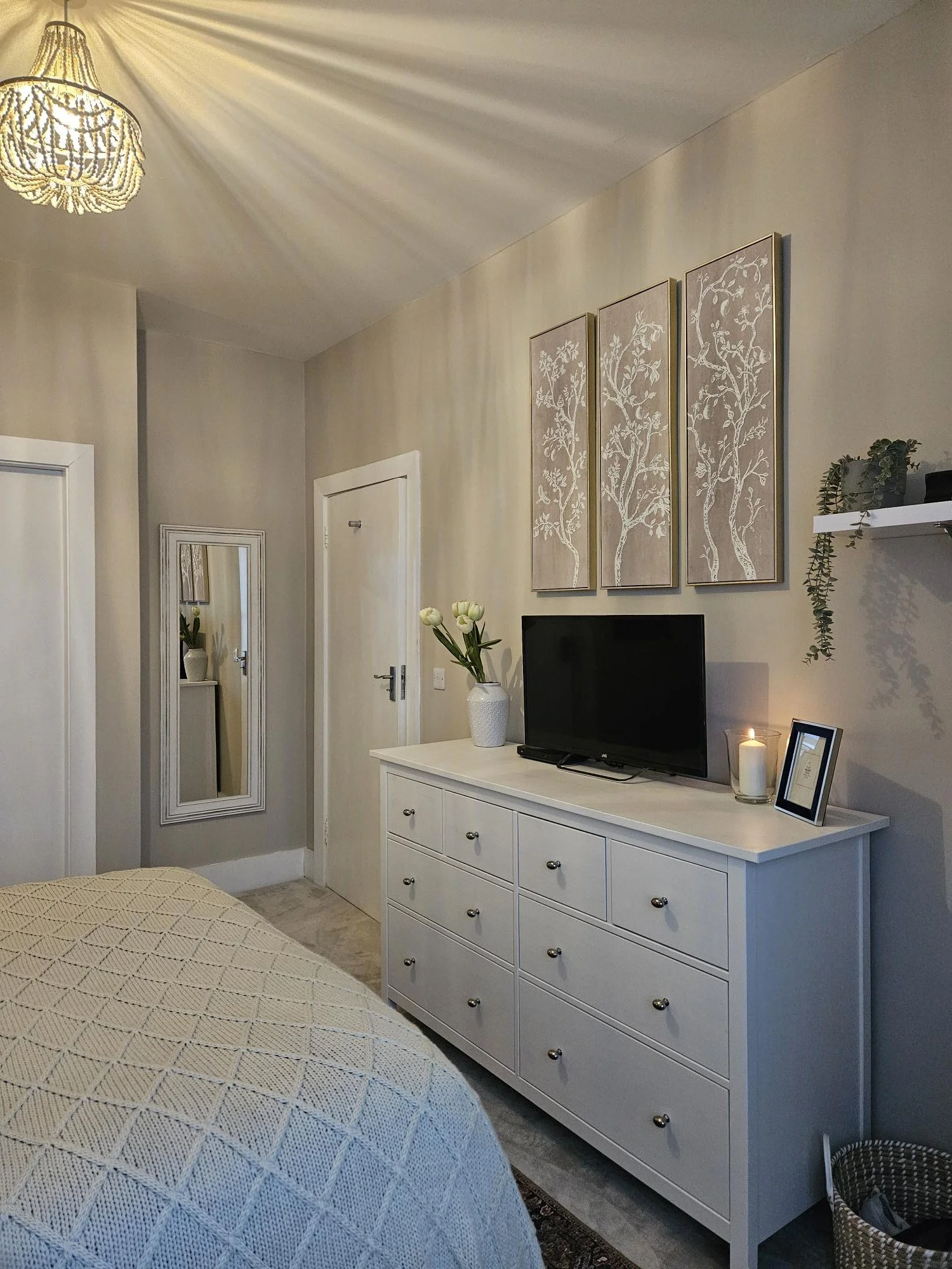 A bedroom dresser with a TV, a candle, framed photo, and a vase of flowers, with wall art and shelf with plants above. By KF Designs