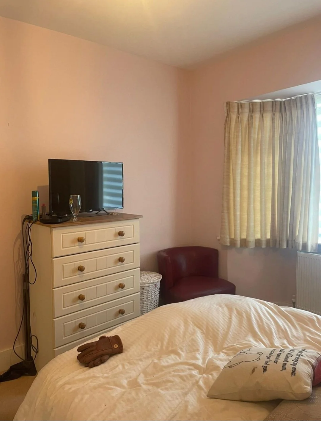 A bedroom with a bed, a dresser with a TV, a pair of gloves on the bed, and a maroon chair near a window with yellow curtains.