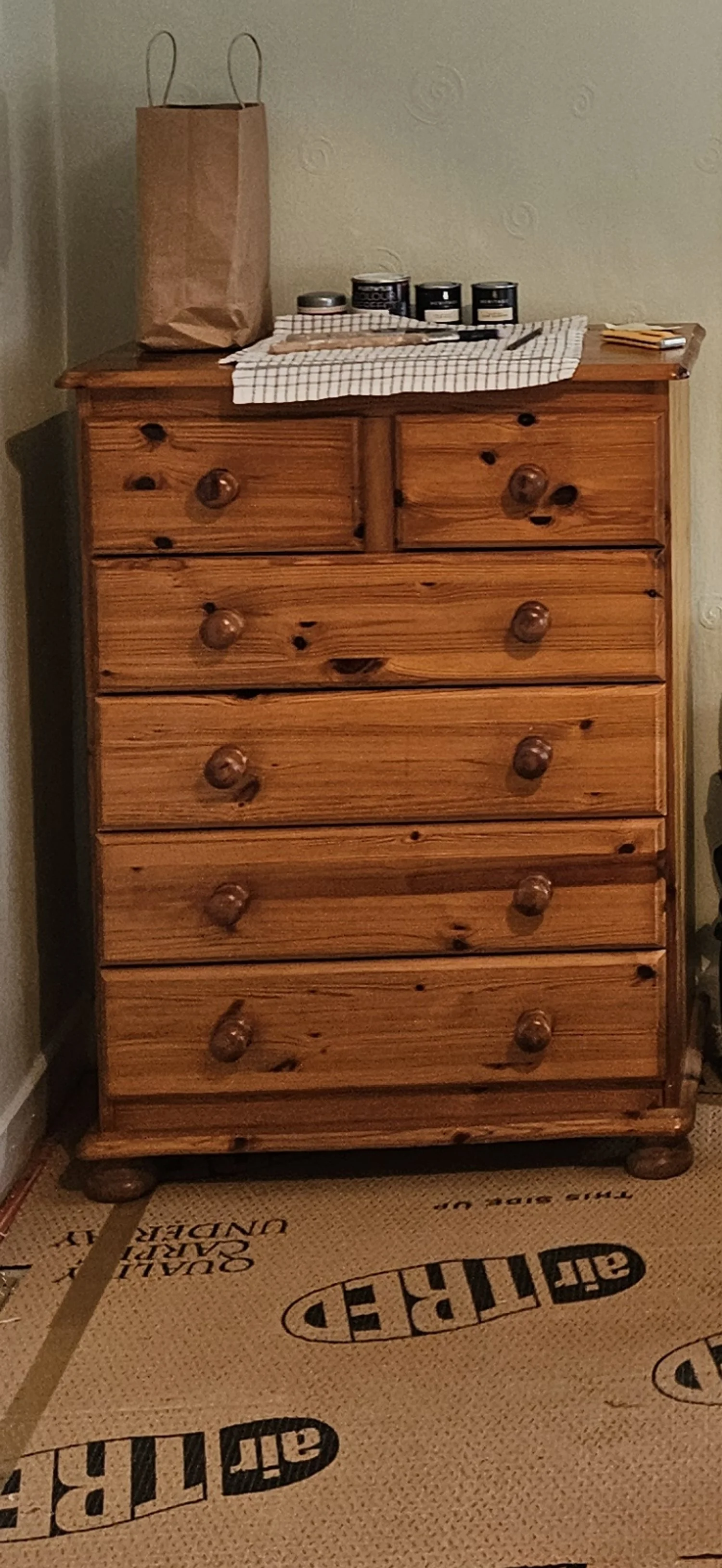 Wooden dresser with seven drawers topped with a paper bag and various cans and items.