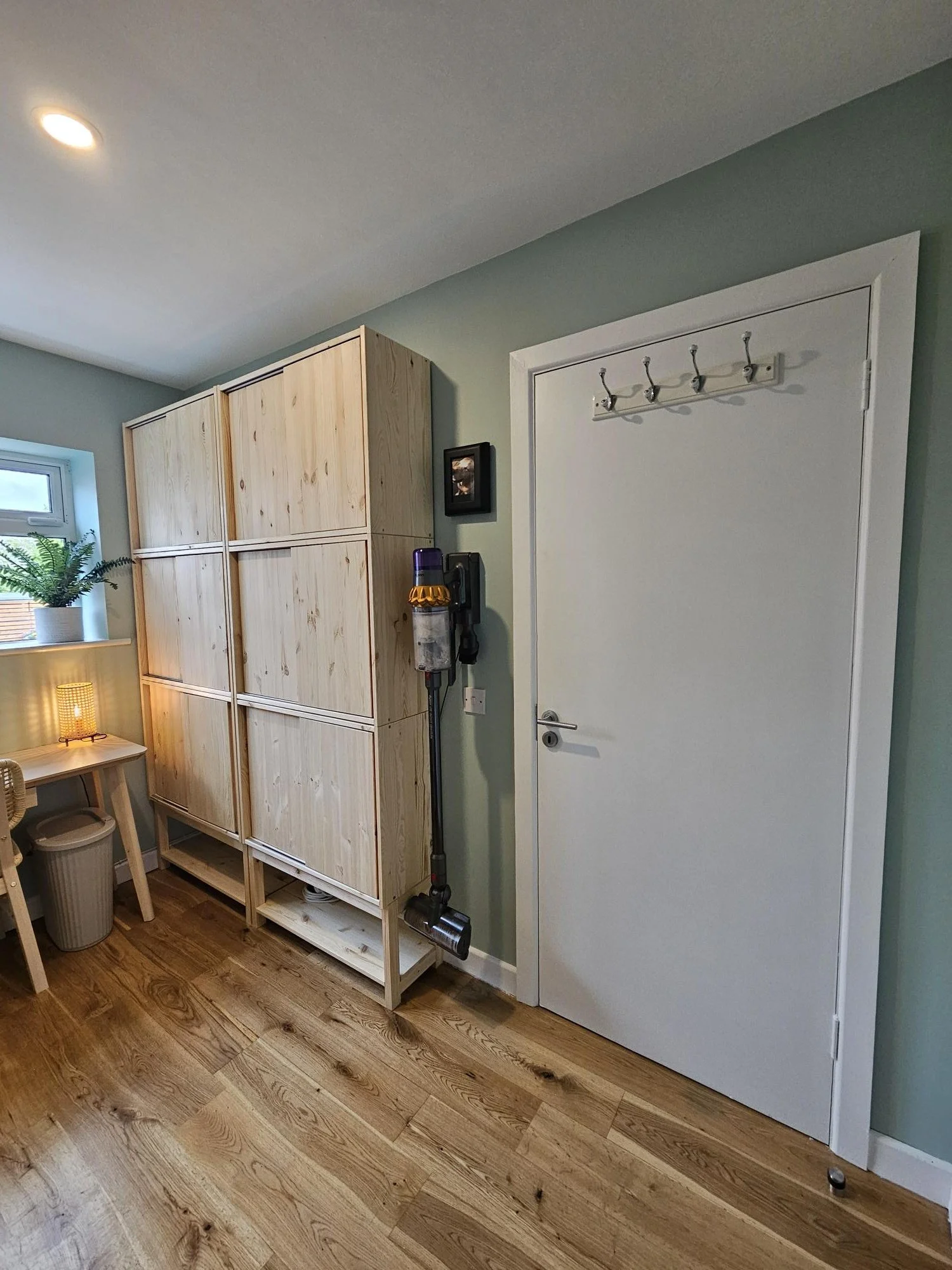 A room with light green walls, hardwood floor, a white door with hooks, a wooden storage unit, a small desk with a lamp, a trash bin, and a window with a potted plant. By KF Designs