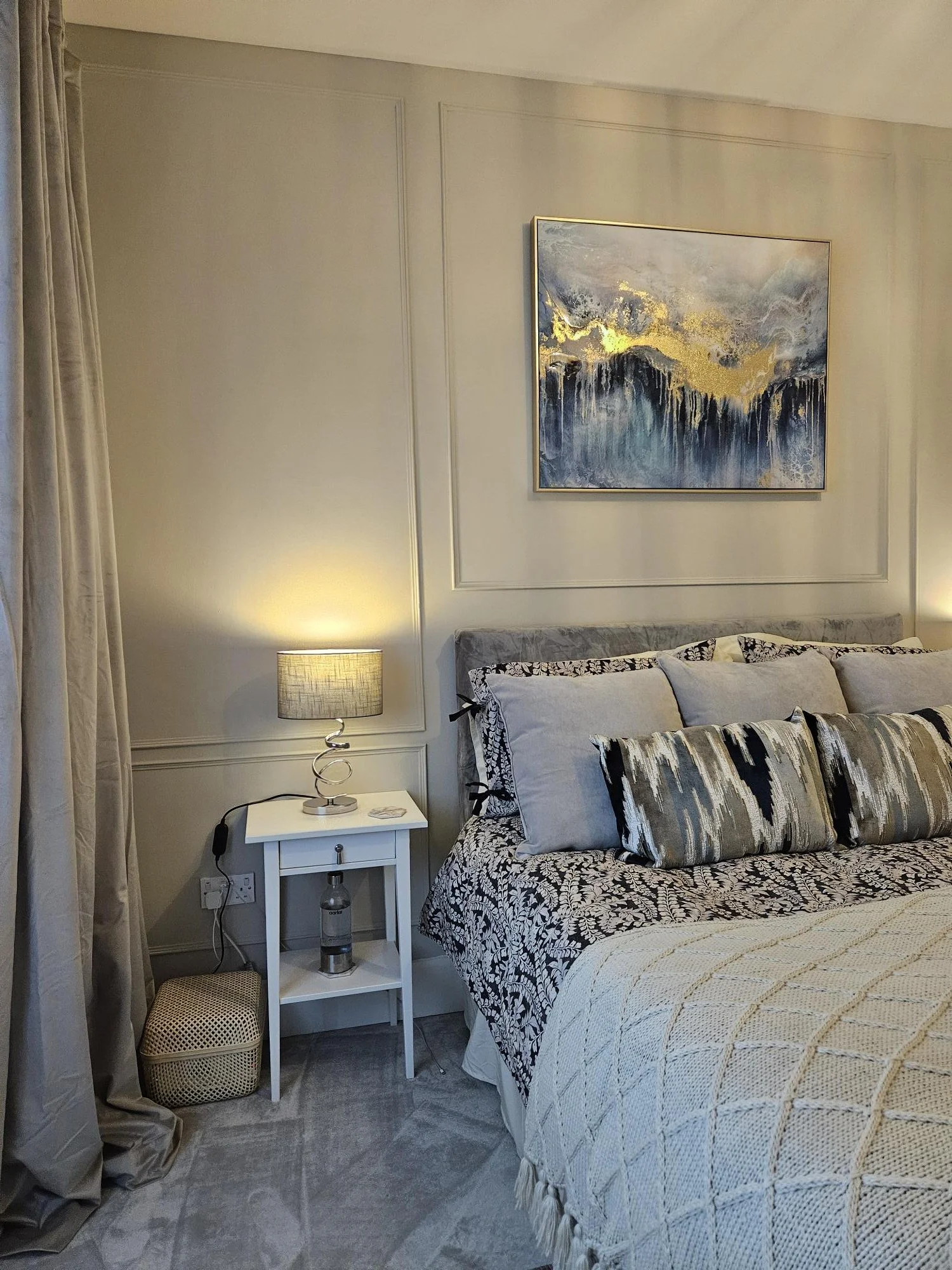 A bedroom corner with a bedside table, lamp, bed with decorative pillows, and an abstract painting on the wall. By KF Designs