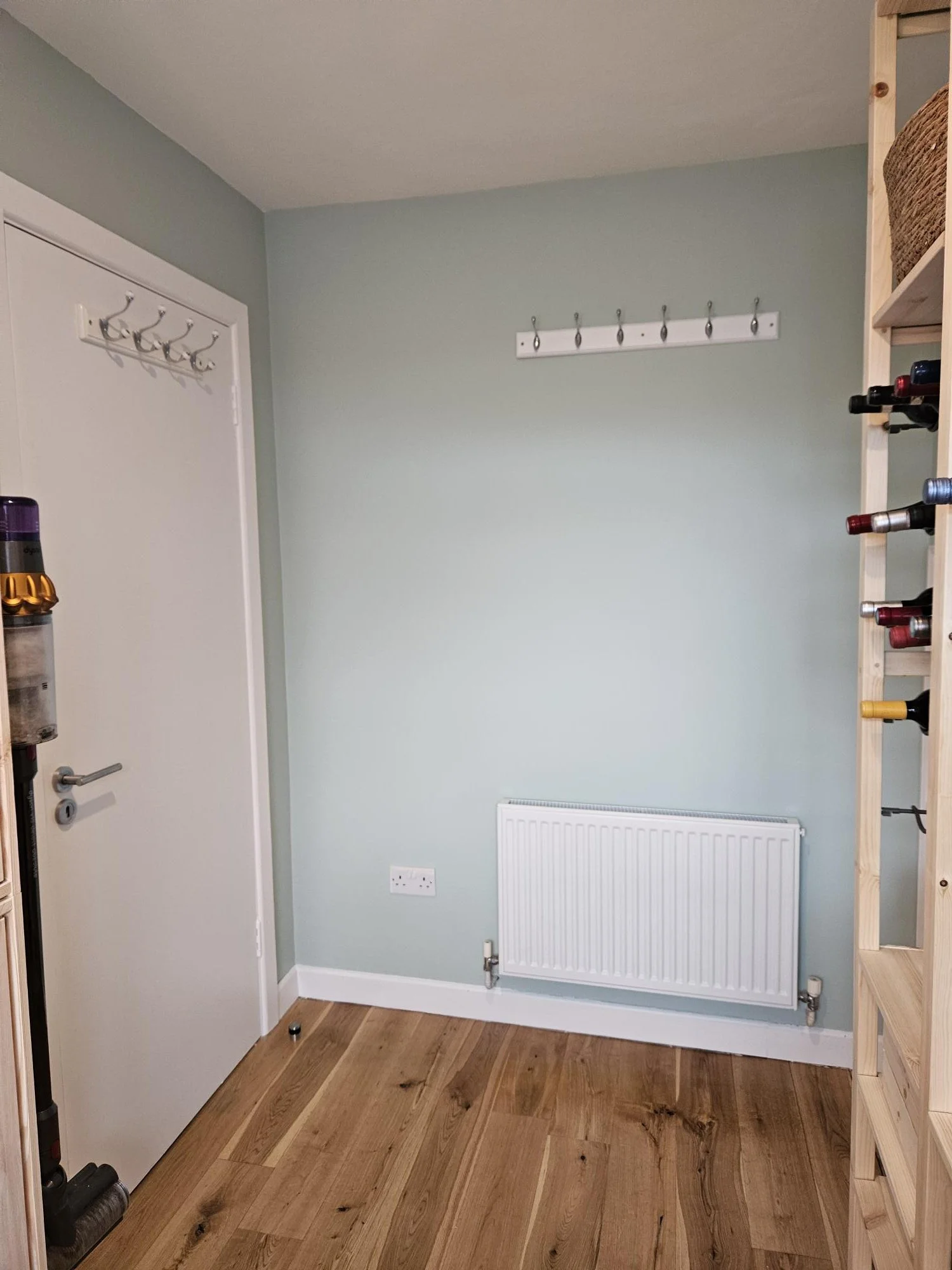 Empty room with a light green wall, a white door, a radiator, a wooden shelf, and hooks on the wall.