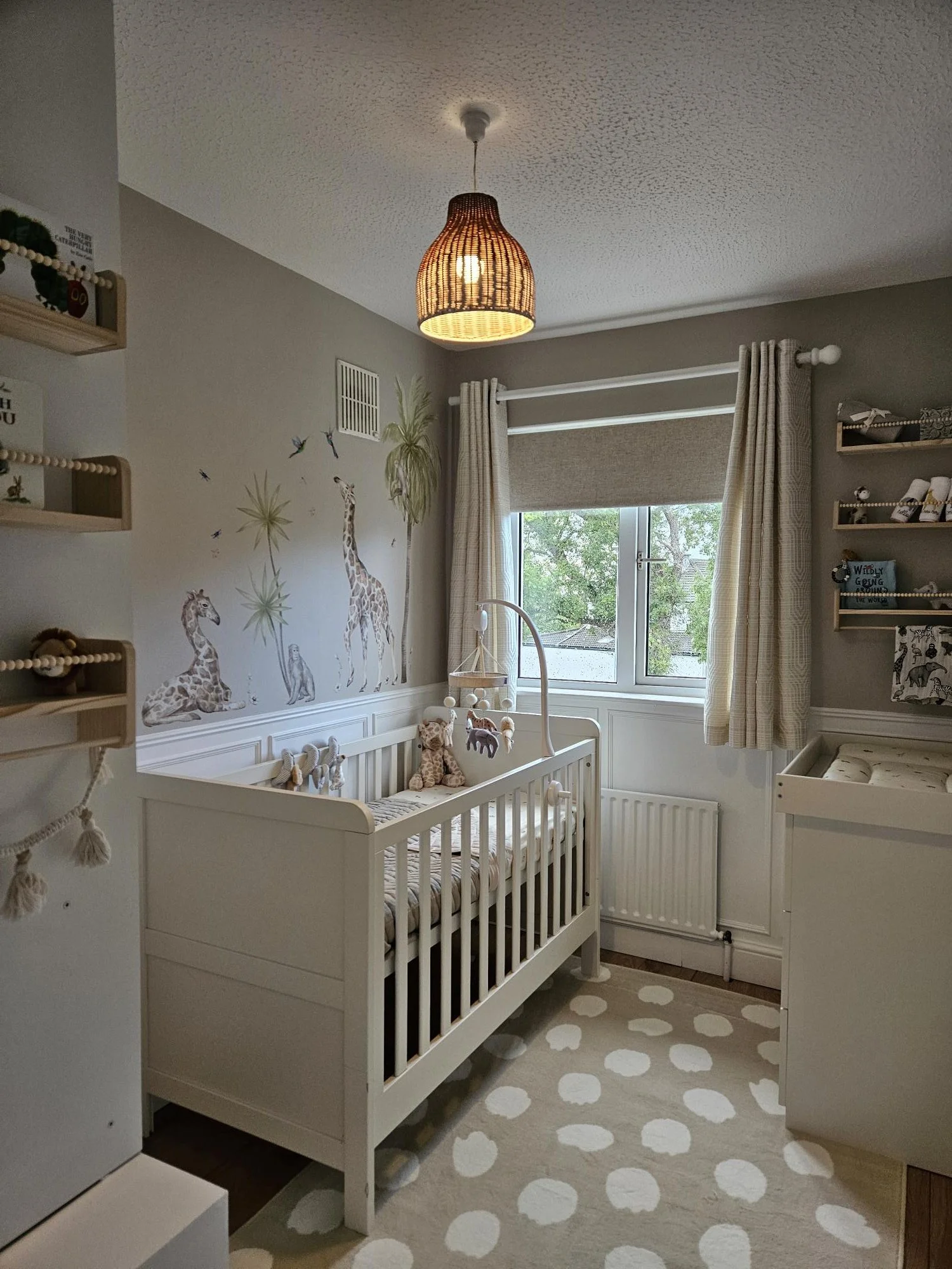 Nursery with a white crib, giraffe wall decal, stuffed animals, window with beige curtains, woven ceiling light, and a patterned rug by KF Designs.