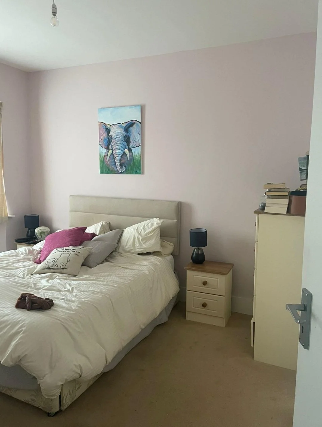 A bedroom with a bed, pillows, bedside tables, lamps, a painting of an elephant, a pile of books, and a window with curtains.