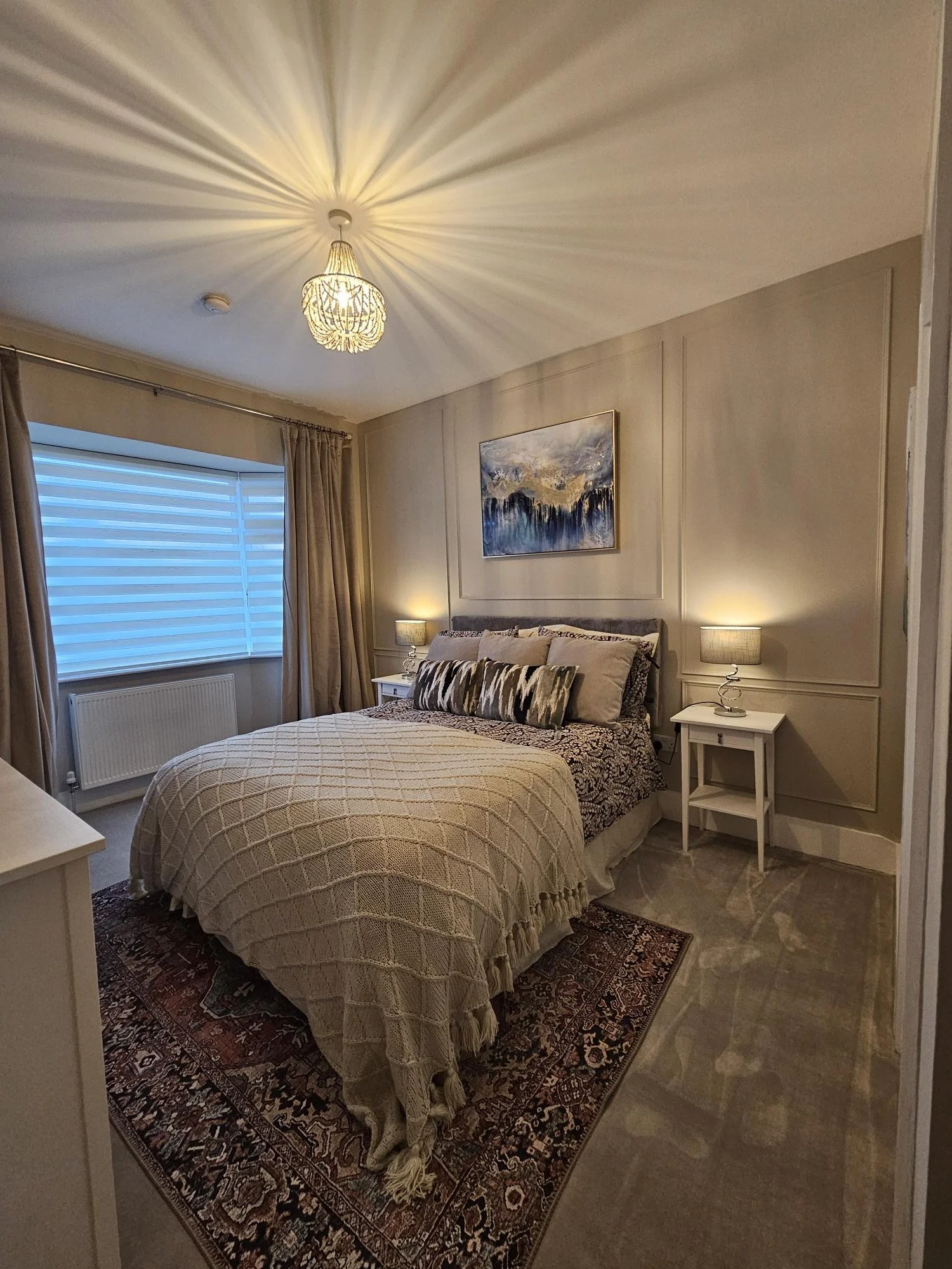 Cozy bedroom with a bed, two nightstands with lamps, a window with blinds and curtains, a painting above the bed, and a decorative rug on the floor. By KF Designs