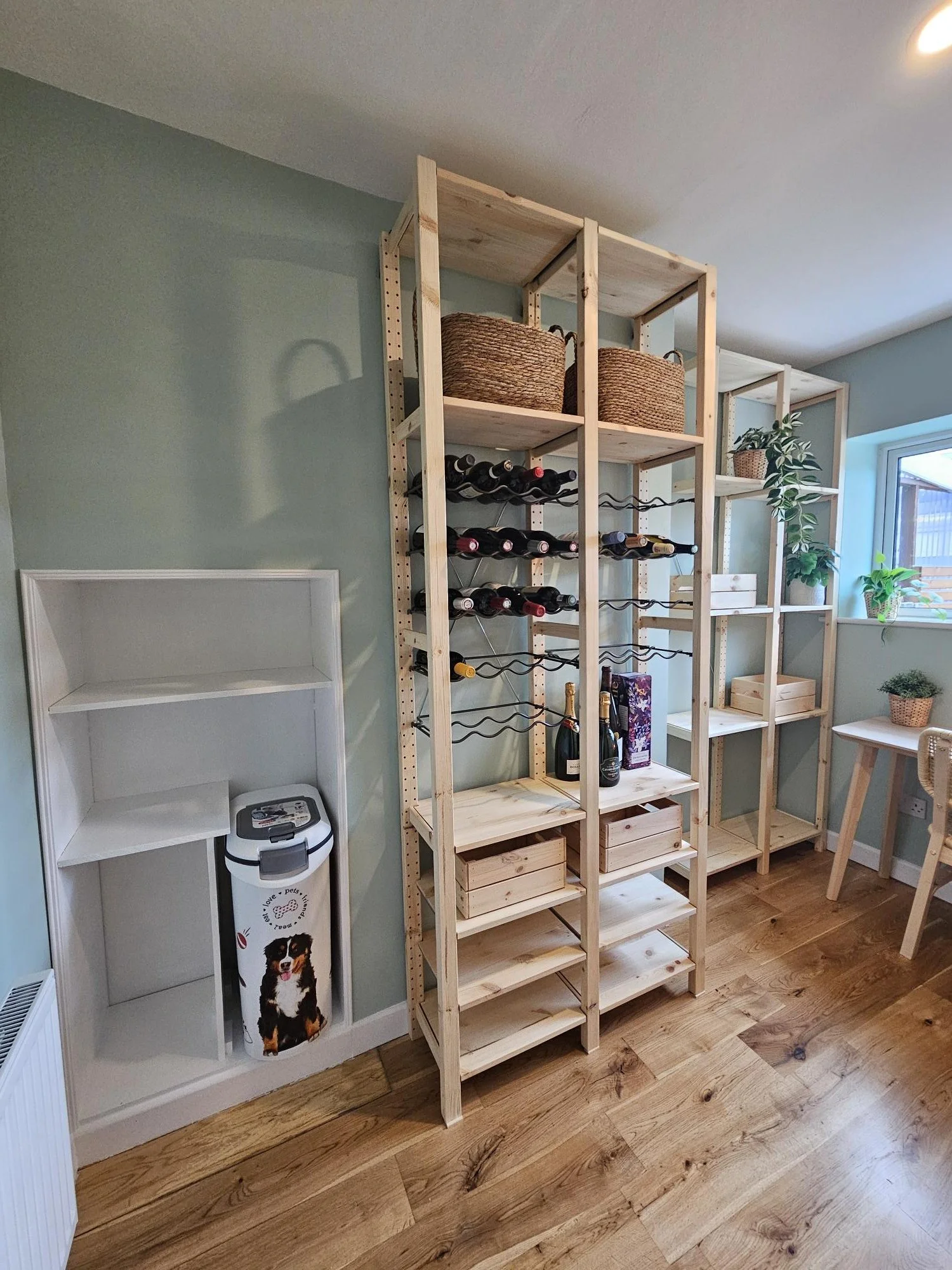 Wooden wine racks and shelves with baskets and plants in a room with wooden flooring and light green walls. By KF Designs