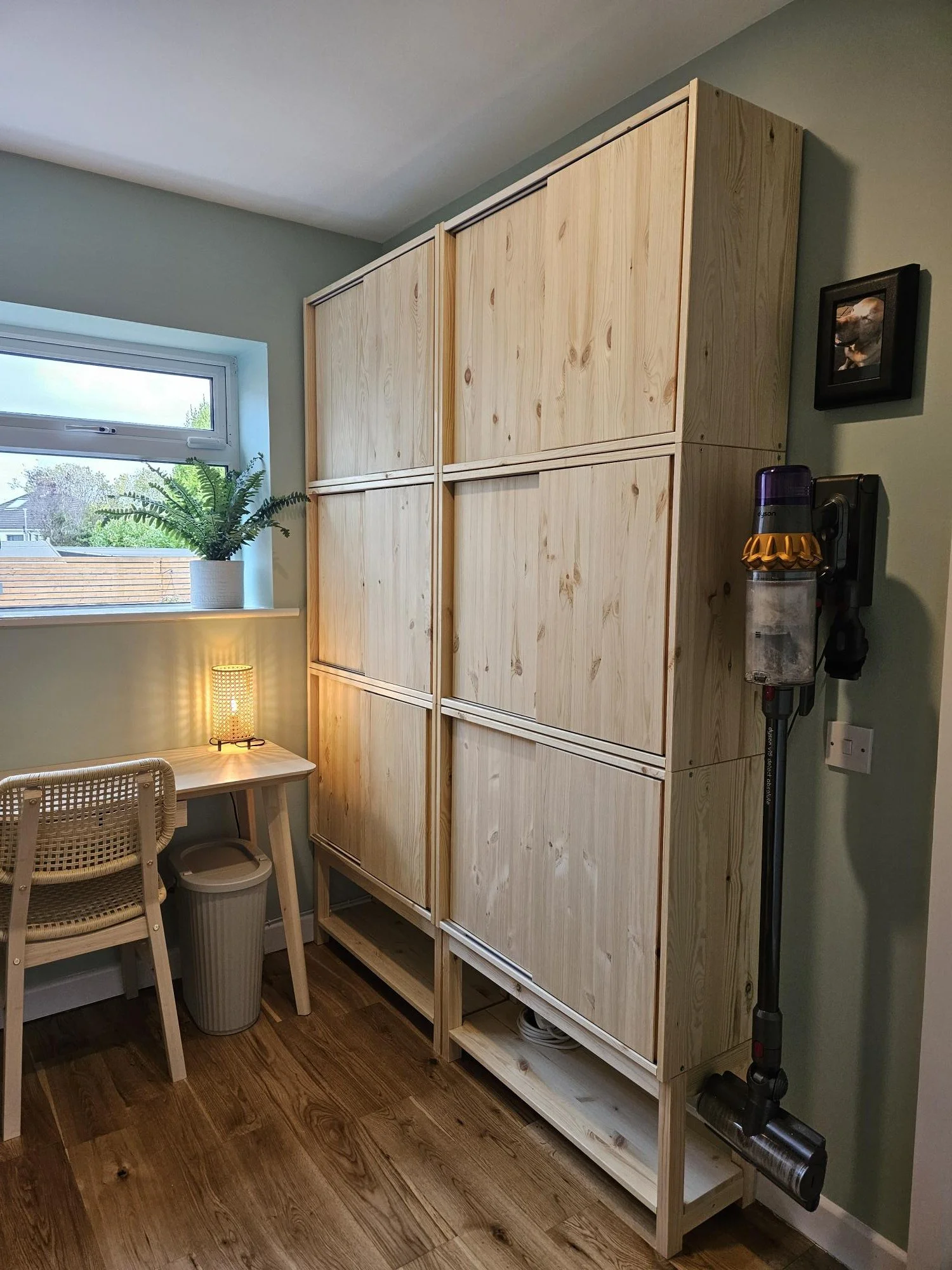Wooden storage cabinet with sliding doors in a room with a window, a small desk with a lamp, and a Dyson vacuum cleaner mounted on the wall. By KF Designs