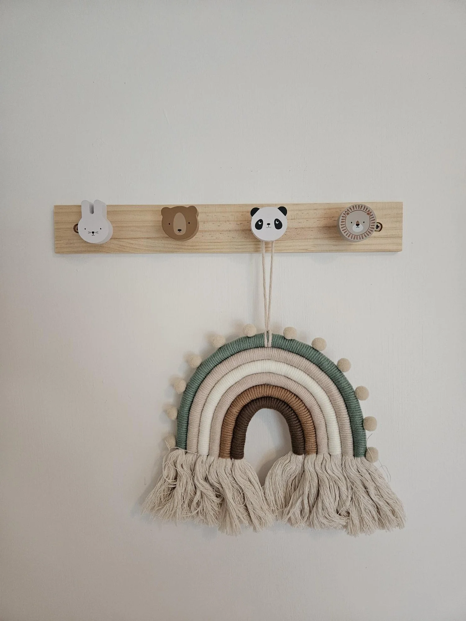 A decorative wall hanging featuring a rainbow made of yarn in earth tones, with white tassels at each end, and a wooden hanger with four animal-shaped clips, including a bunny, a bear, a panda, and a lion.