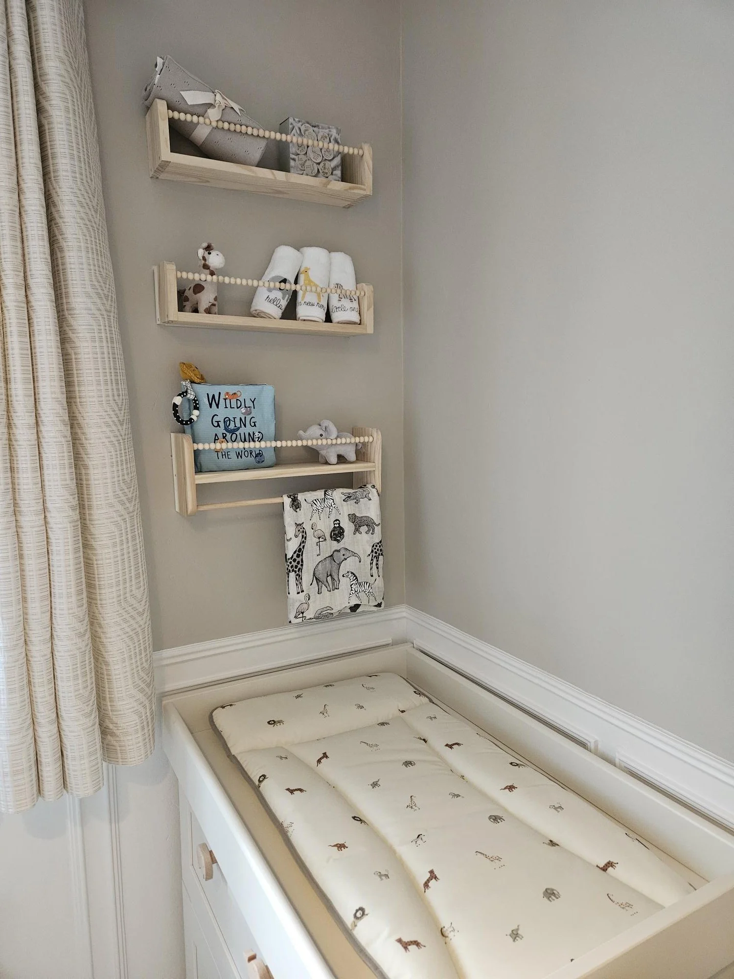 Corner of a nursery with three wooden shelves decorated with plush toys and towels, a changing table with animal-themed padding, and beige curtains on a window. By KF Designs