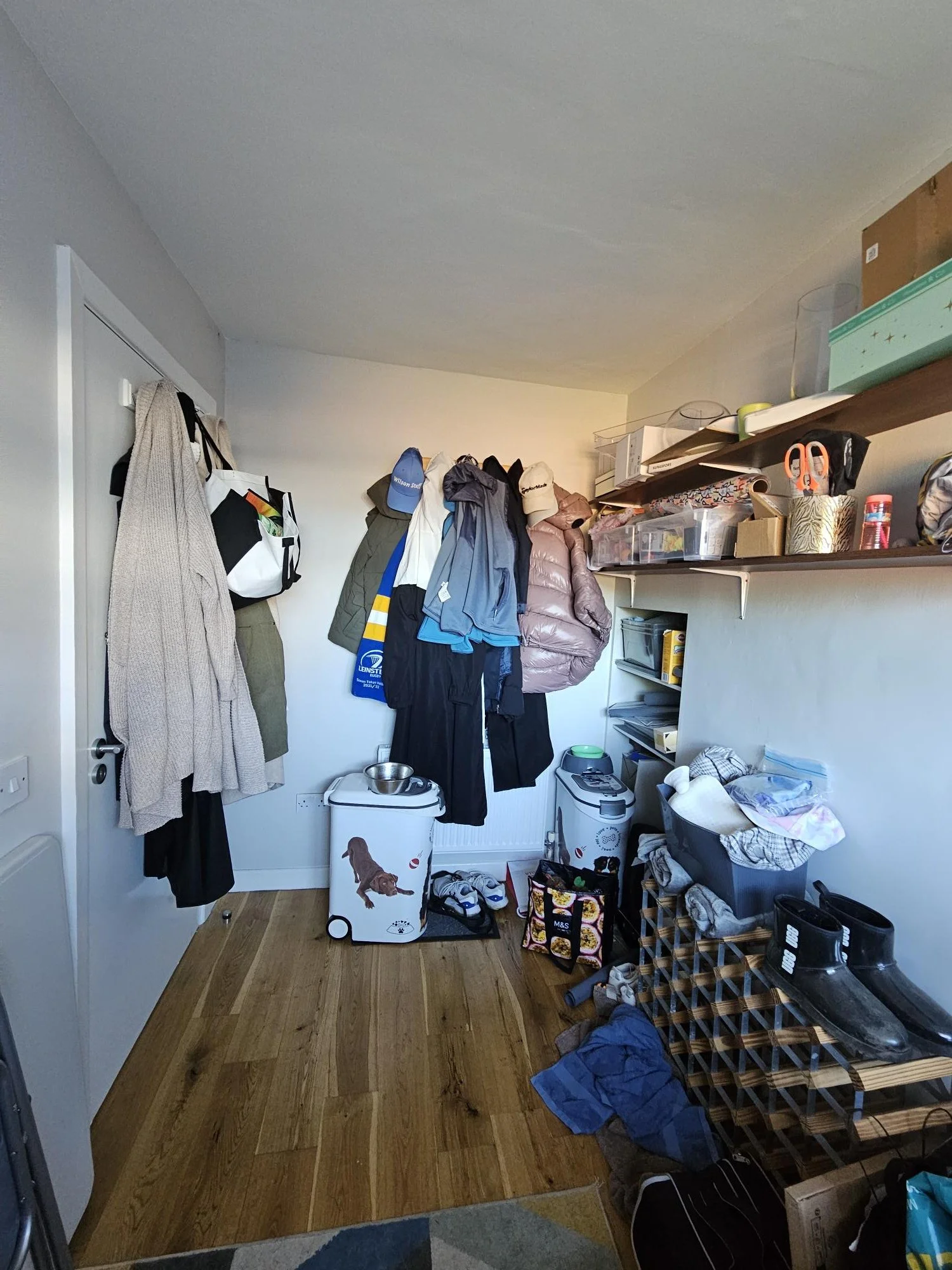 A cluttered room with hanging clothes, shelves with boxes and items, and shoes on a wooden shoe rack.