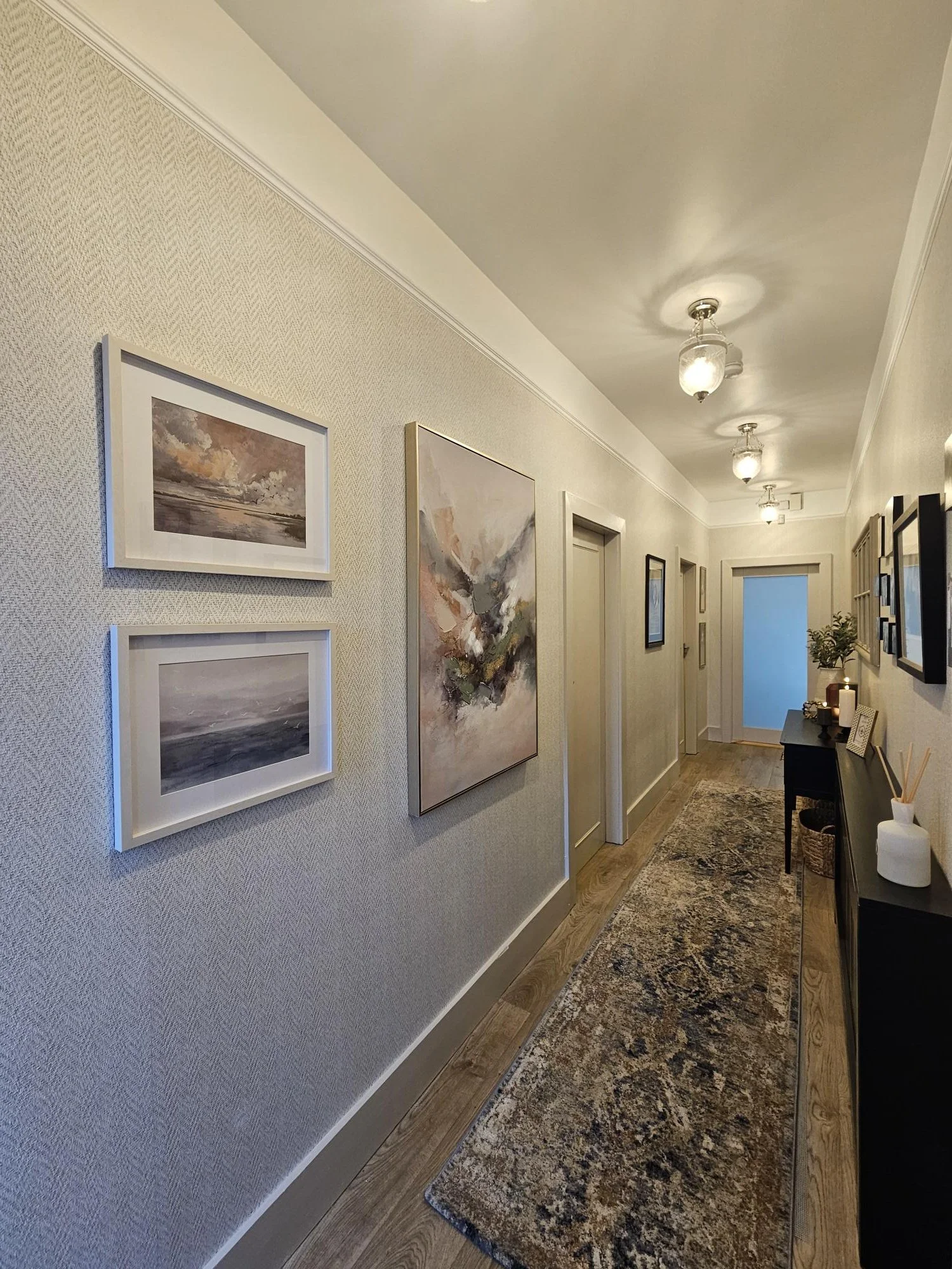 A hallway with several ceiling lights, framed artwork on the wall, a black console table with decorative items, and a patterned rug on wooden flooring. By KF Designs