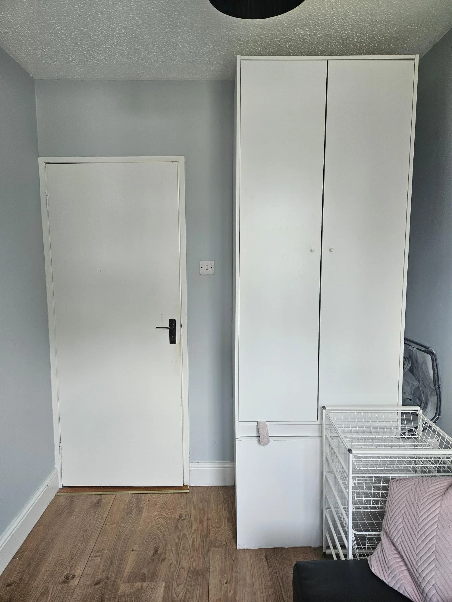 A white door with a black handle, a white wardrobe, a white wire basket, a pink pillow on a black chair, and a folded drying rack against a gray wall and wooden floor.