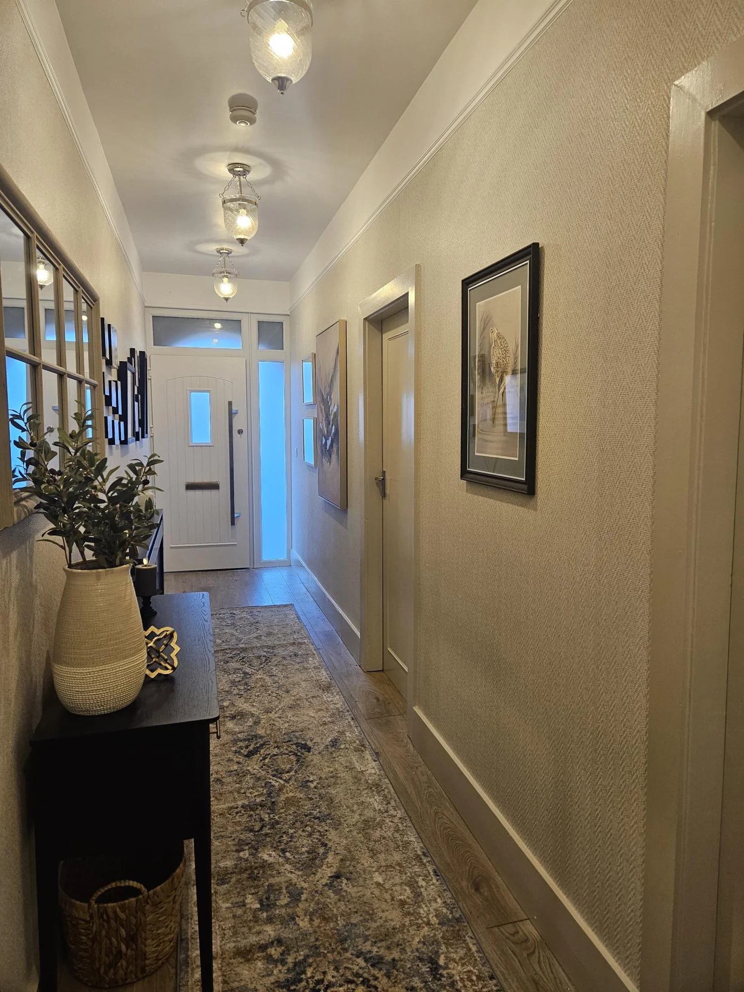 Hallway with a front door at the back, decorated with framed artwork and a console table with a potted plant, rug, and baskets, illuminated by ceiling lights. By KF Designs