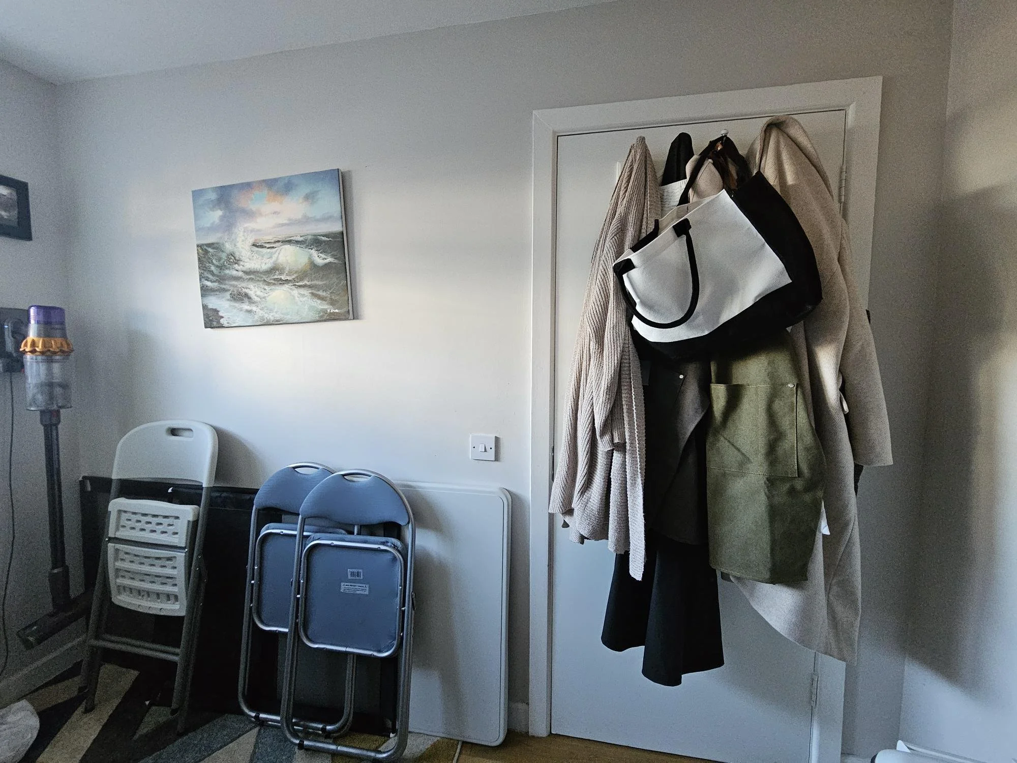 Coat rack with coats, bags, and umbrellas next to a closed door in a room. Folded chairs, a small table, and an artwork of ocean waves on the wall.