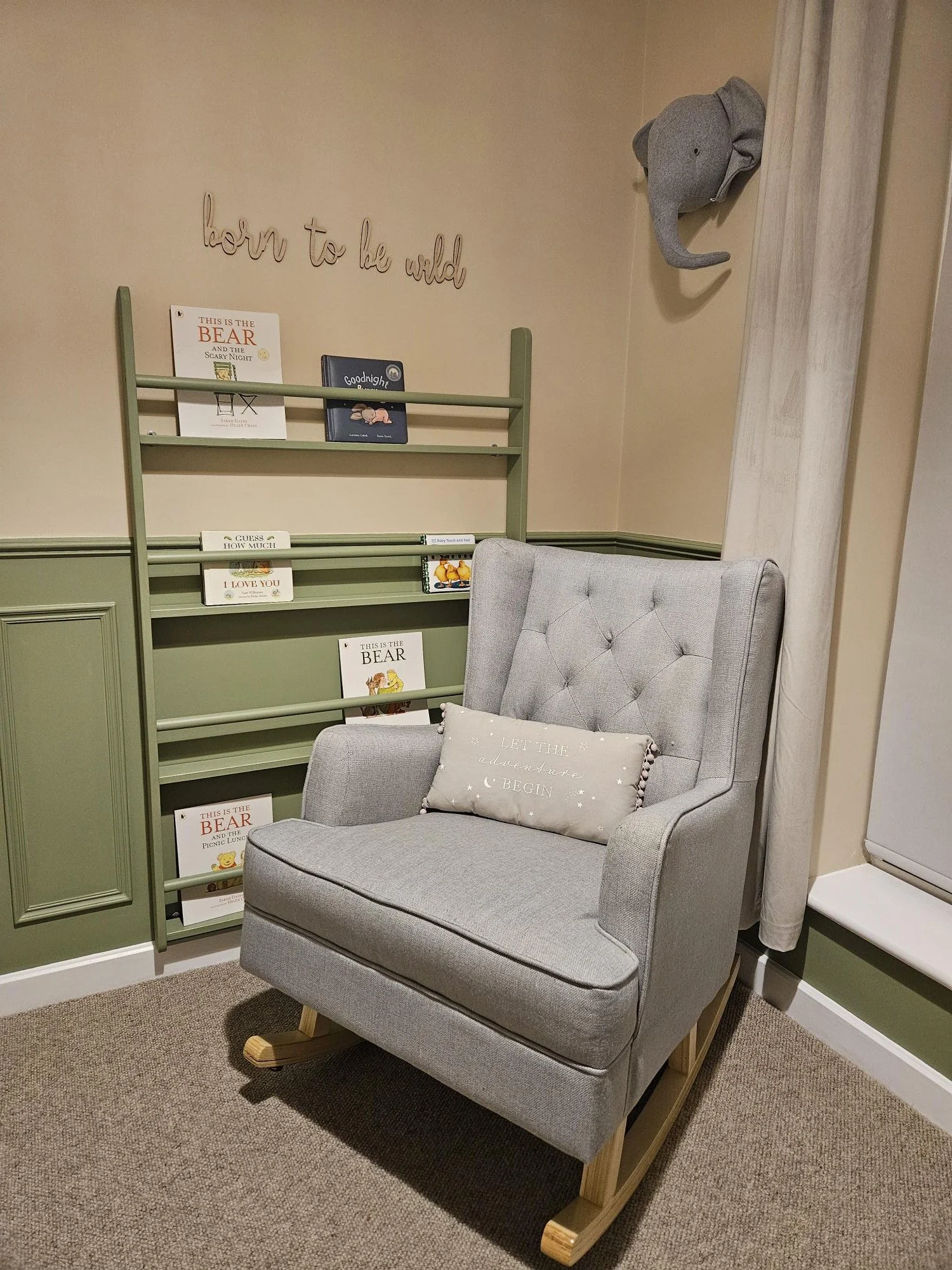 A cozy reading nook with a light grey rocking armchair and a decorative pillow. A green bookshelf with children’s books and a wall sign that reads 'born to be wild' are in the background by KF Designs.