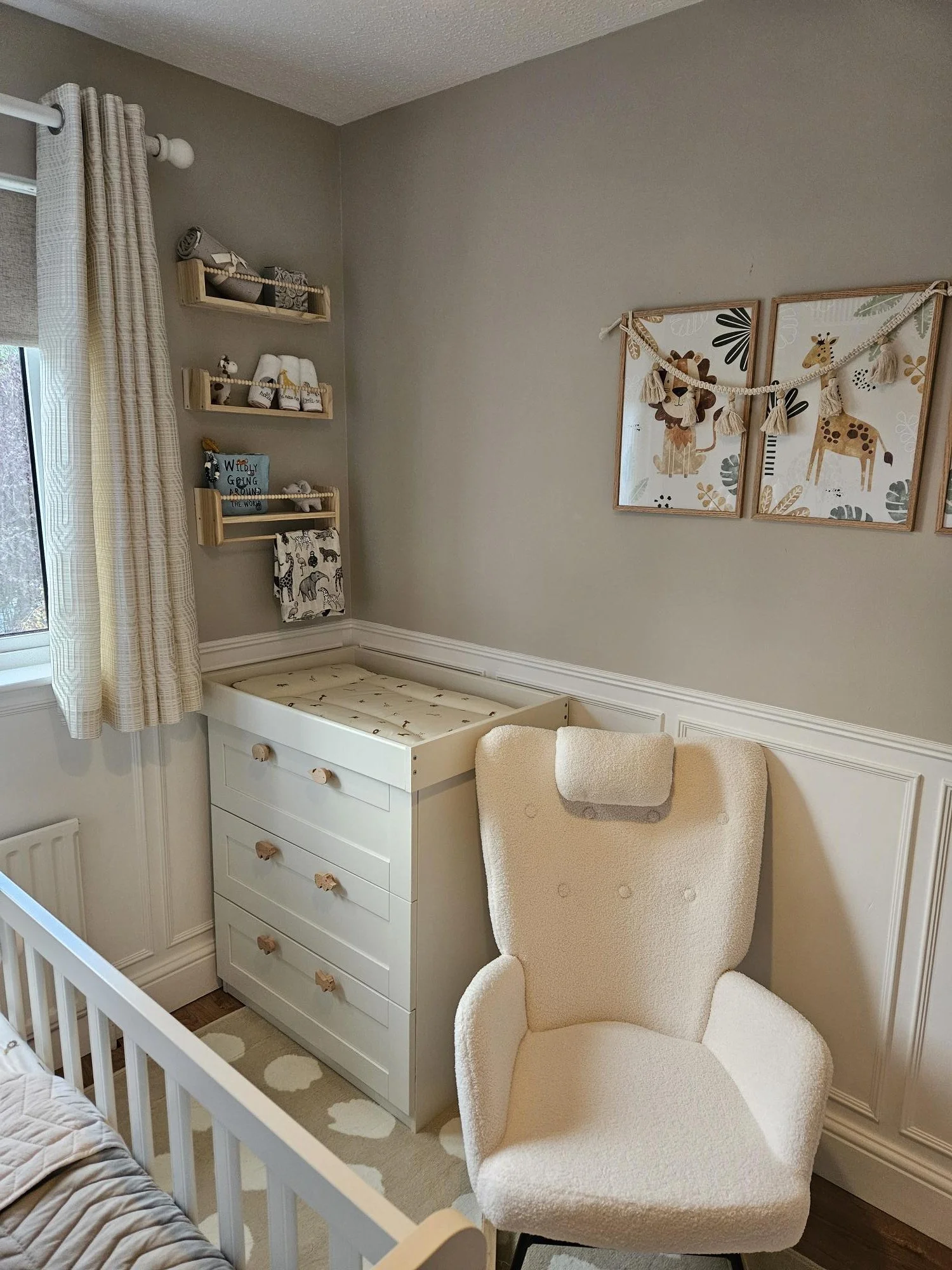 A cozy nursery corner with a white changing table, a cream armchair with a headrest, wall-mounted shelves with baby items, and animal-themed wall art with garland decorations. By KF Designs