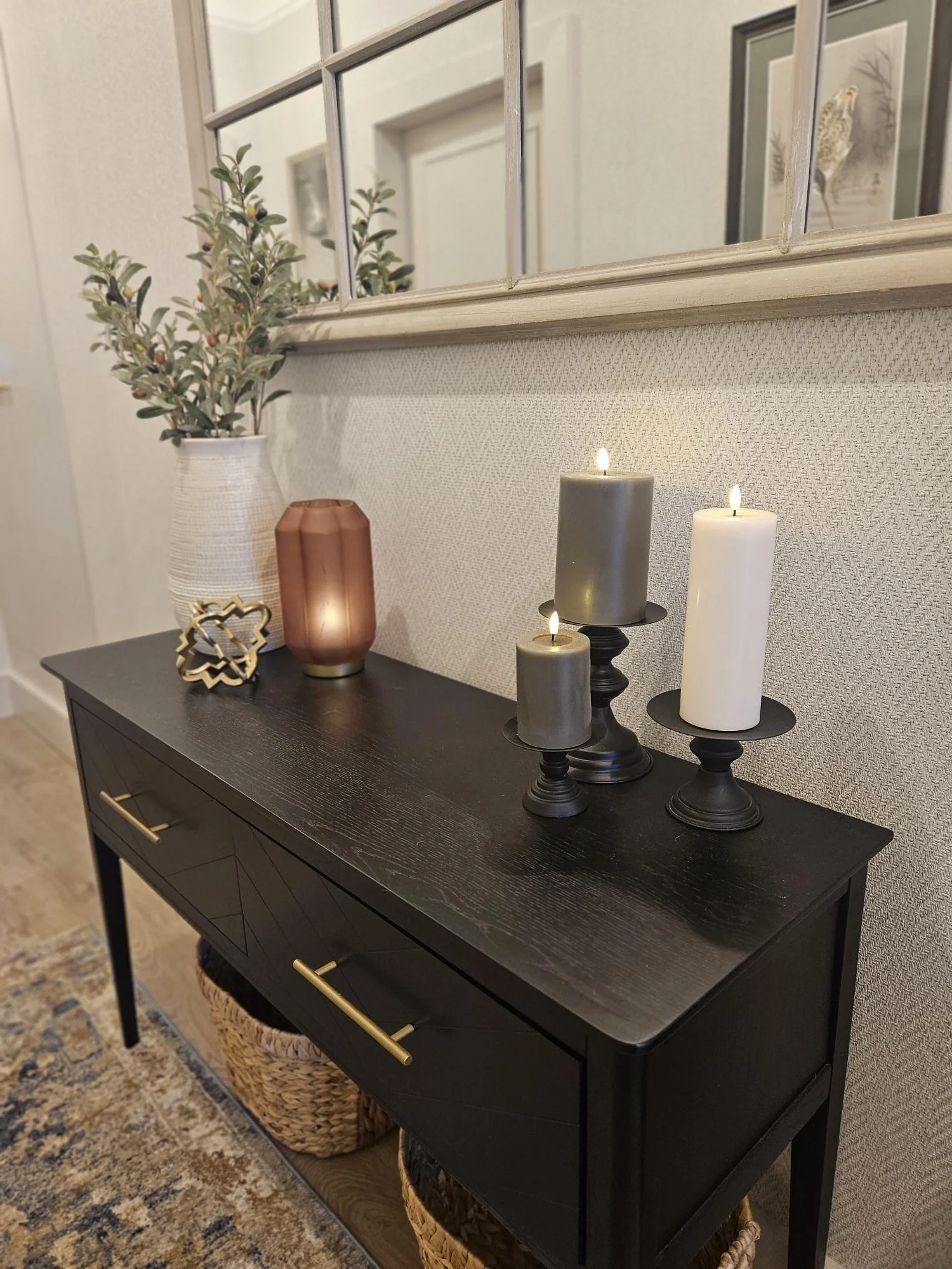 A black console table with gold handles holds a potted plant, a decorative candle holder, and three lit candles in grey, dark grey, and white. Baskets are stored underneath the table, and a mirror hangs above with framed artwork reflected in it. By K
