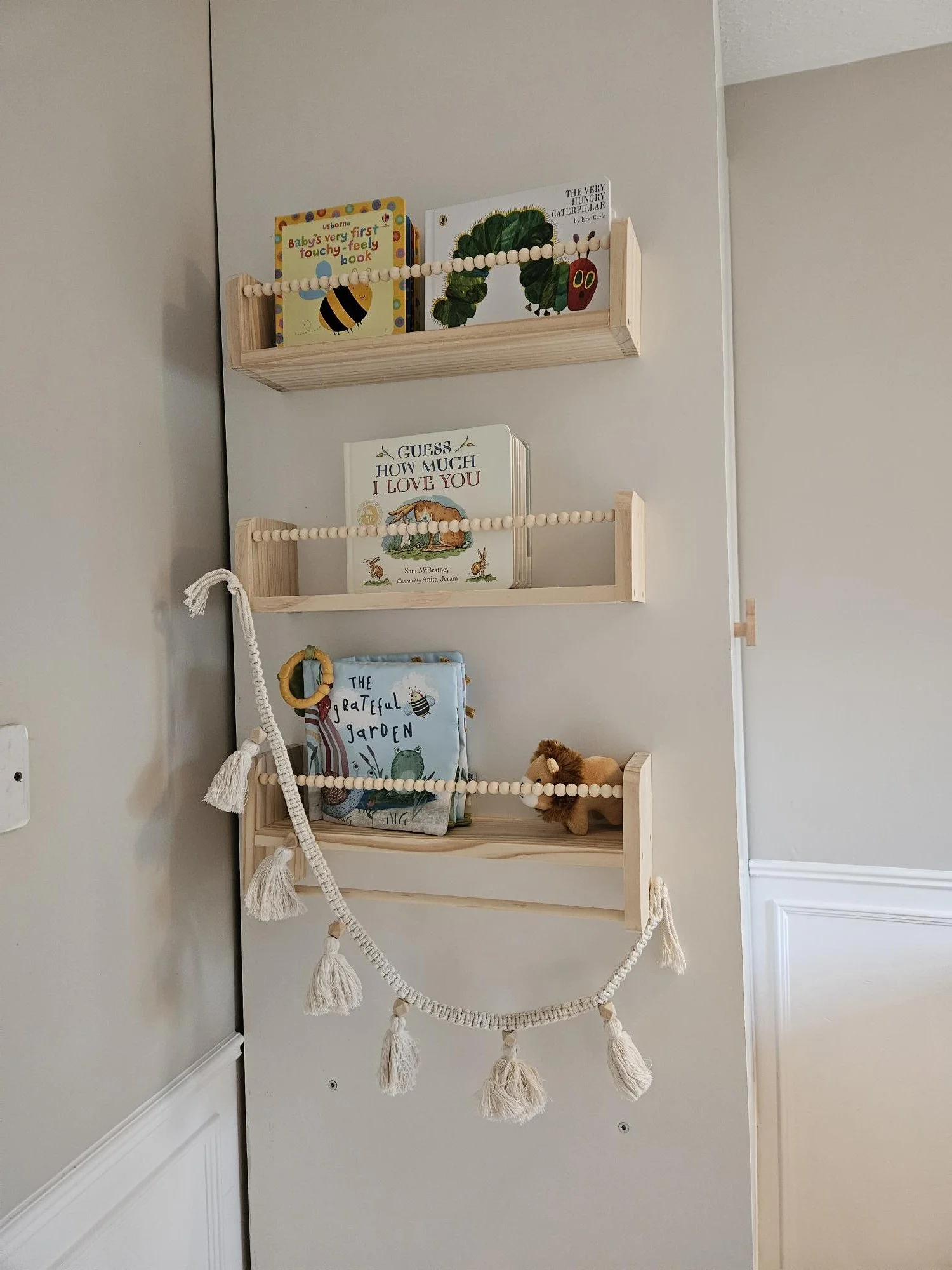 Wall-mounted wooden shelves holding children's books, a plush lion toy, and a decorative tassel garland. By KF Designs