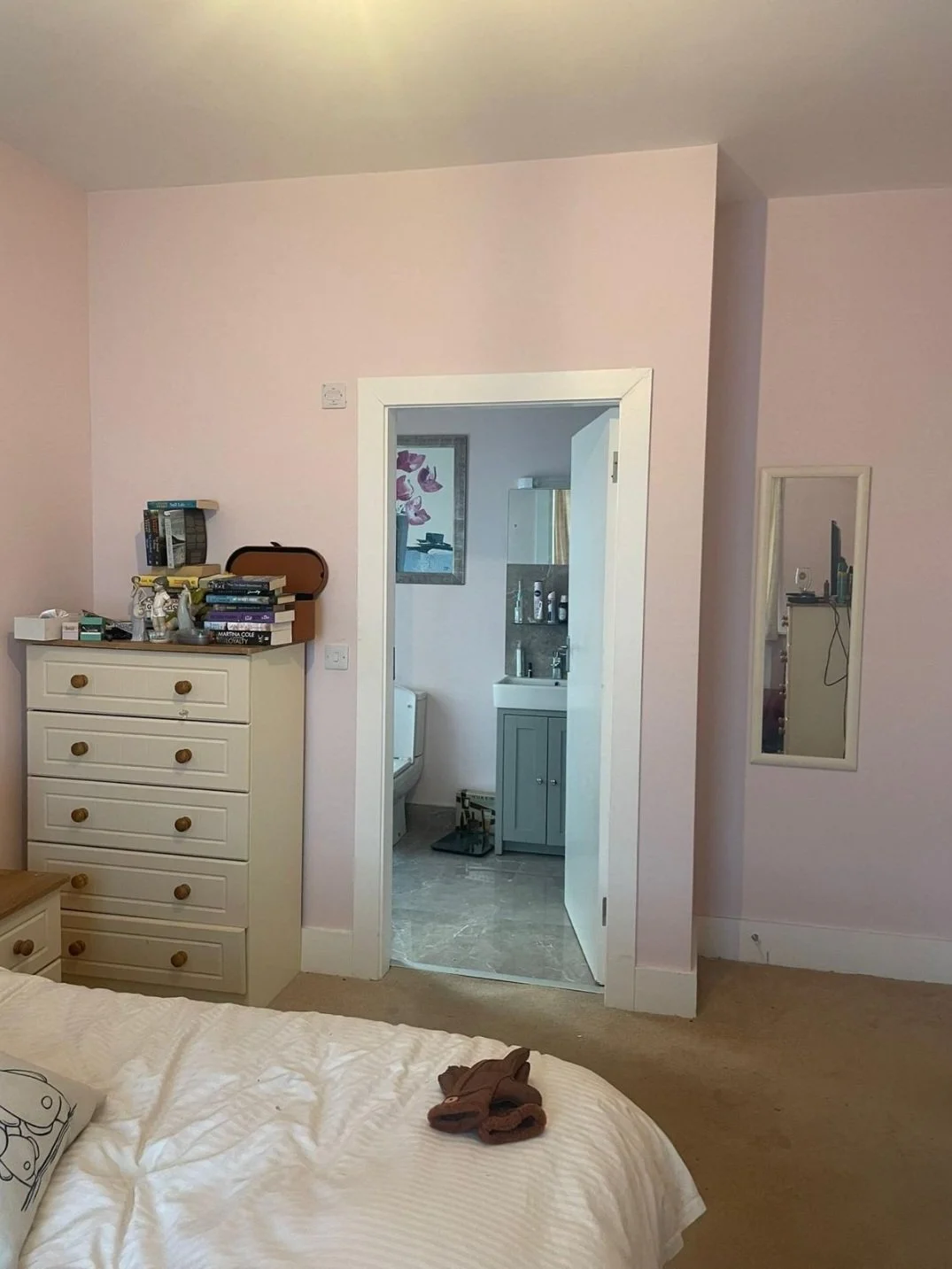 A bedroom with a dresser on the left, a bed at the bottom, and an open door leading to a bathroom with a toilet and sink. A mirror is mounted on the right wall, reflecting part of the room.