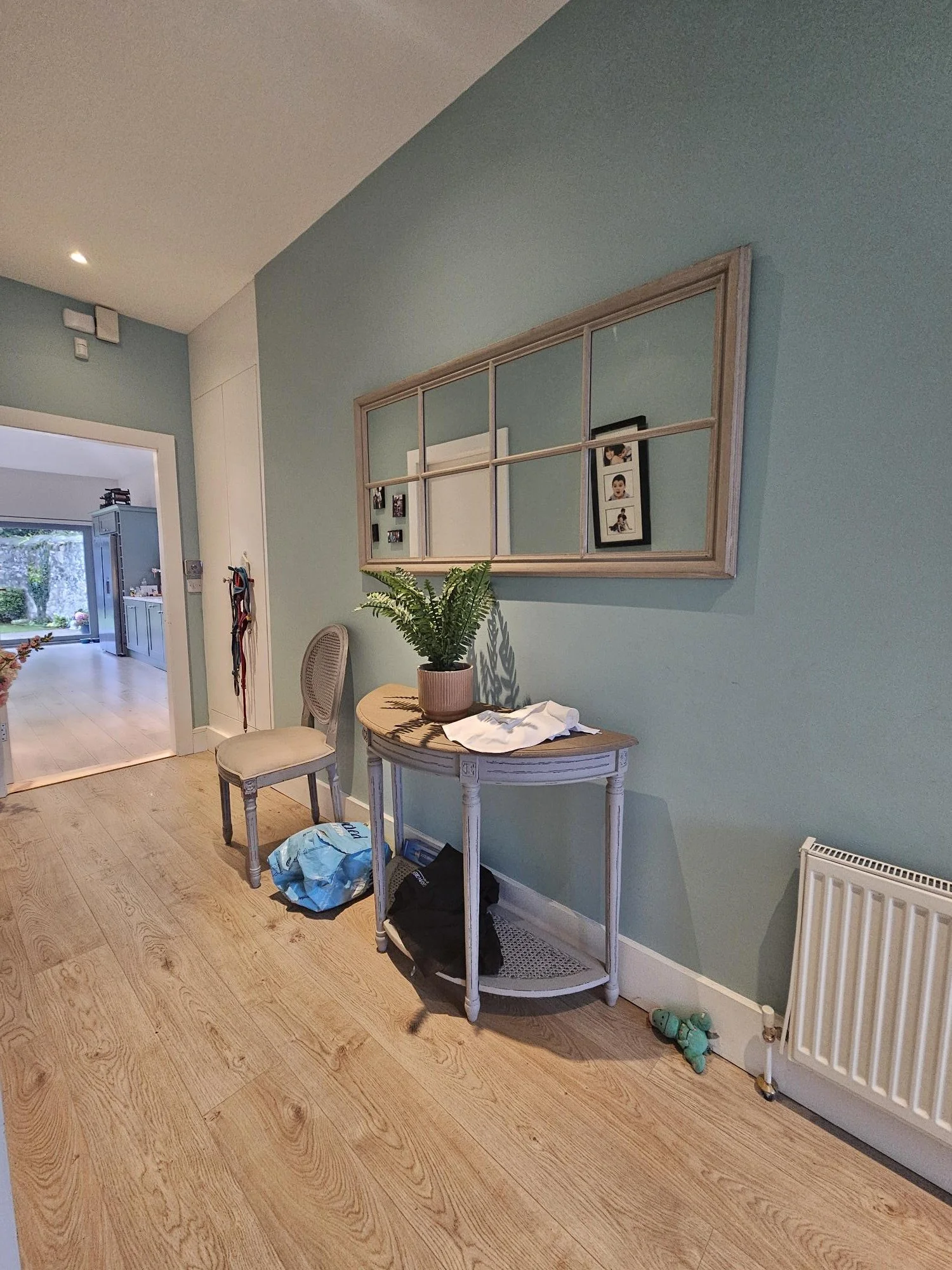 Interior of a home entryway with a vintage console table, potted plant, and a mirror on the wall, leading to a kitchen and living area.