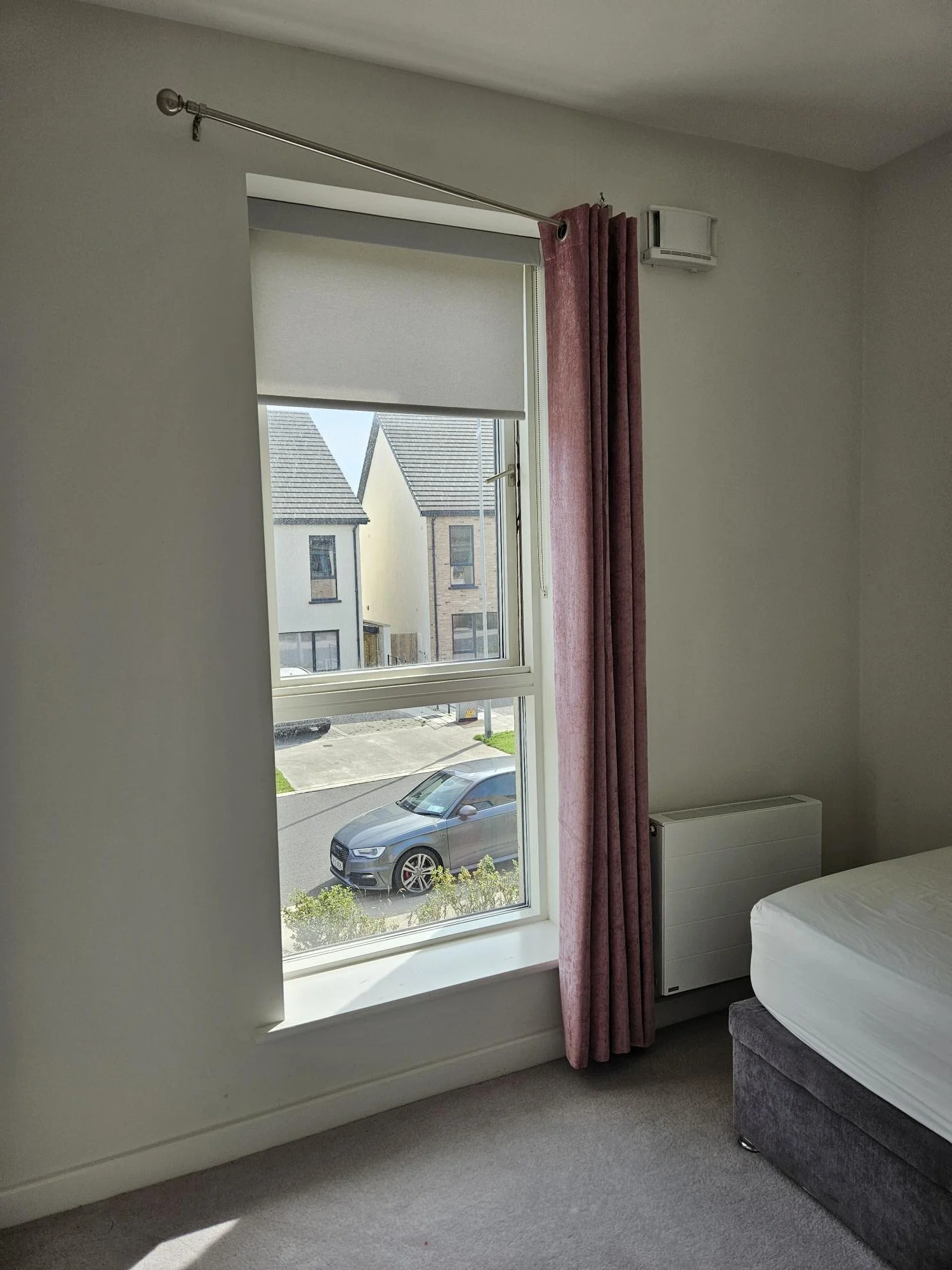 Interior of a bedroom with a window, pink curtains, a white modern heater, part of a bed with a white sheet, and a view of parked cars and neighboring houses outside.