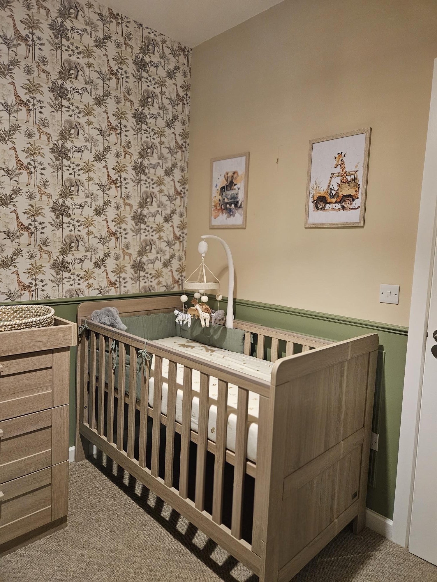 Wooden baby crib with stuffed animals and mobile, in a nursery with animal-themed wallpaper and framed animal art by KF Designs