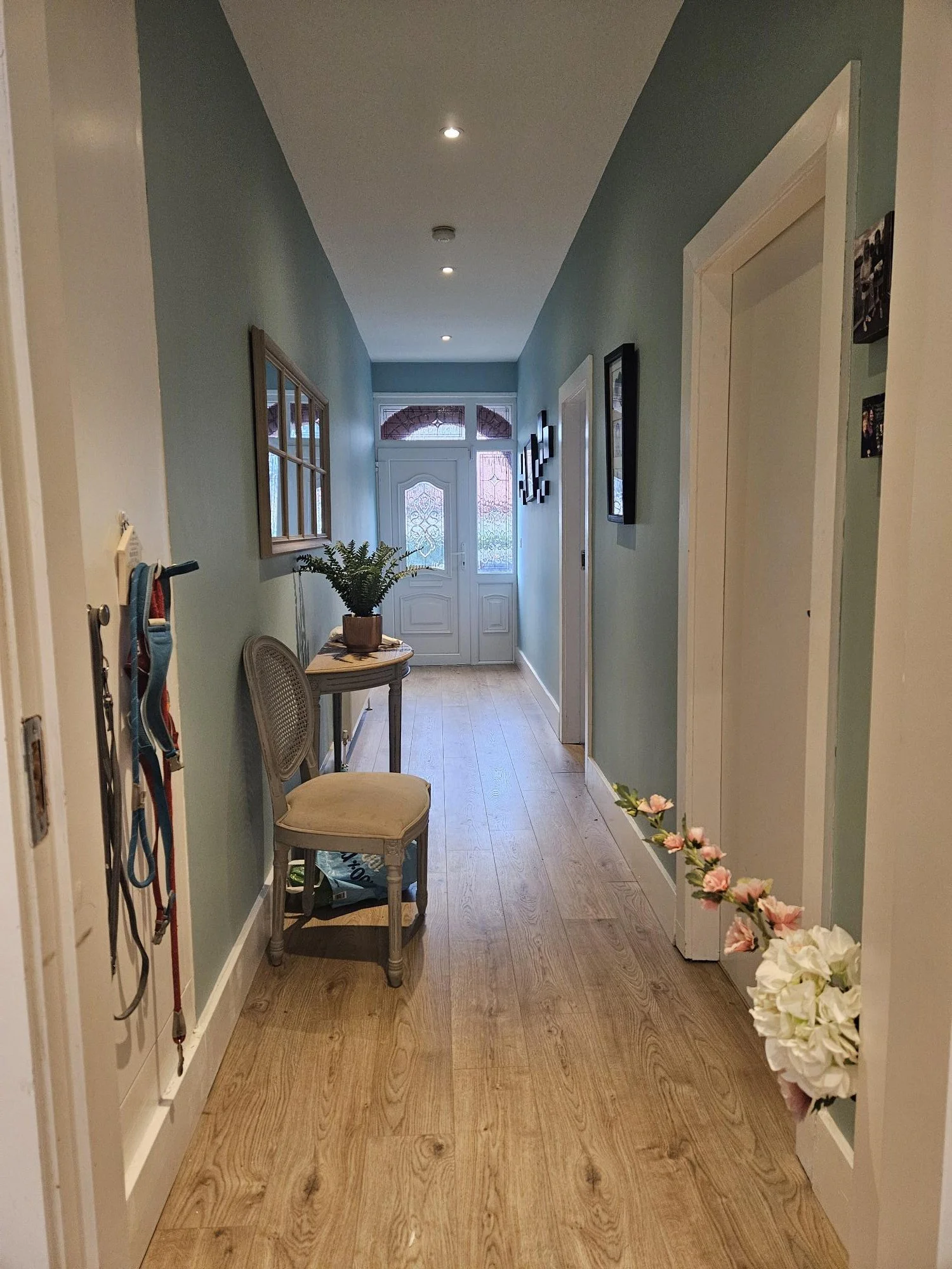 A hallway with light green walls, hardwood floor, and white door at the end. Decor includes framed pictures, a chair with a potted plant, and a floral arrangement.