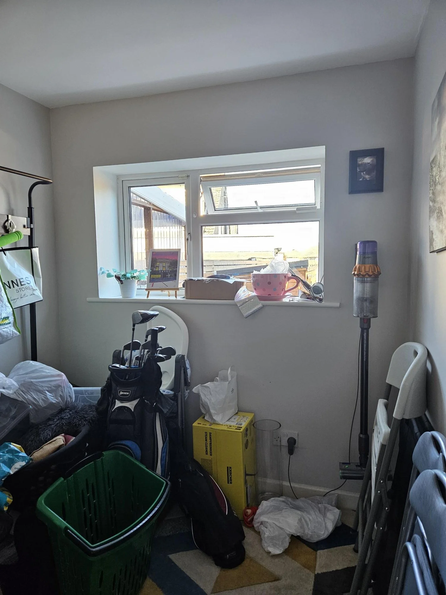 A cluttered room with a window, a vacuum cleaner, a golf bag, a green laundry basket, and various scattered items including tissue paper and boxes.
