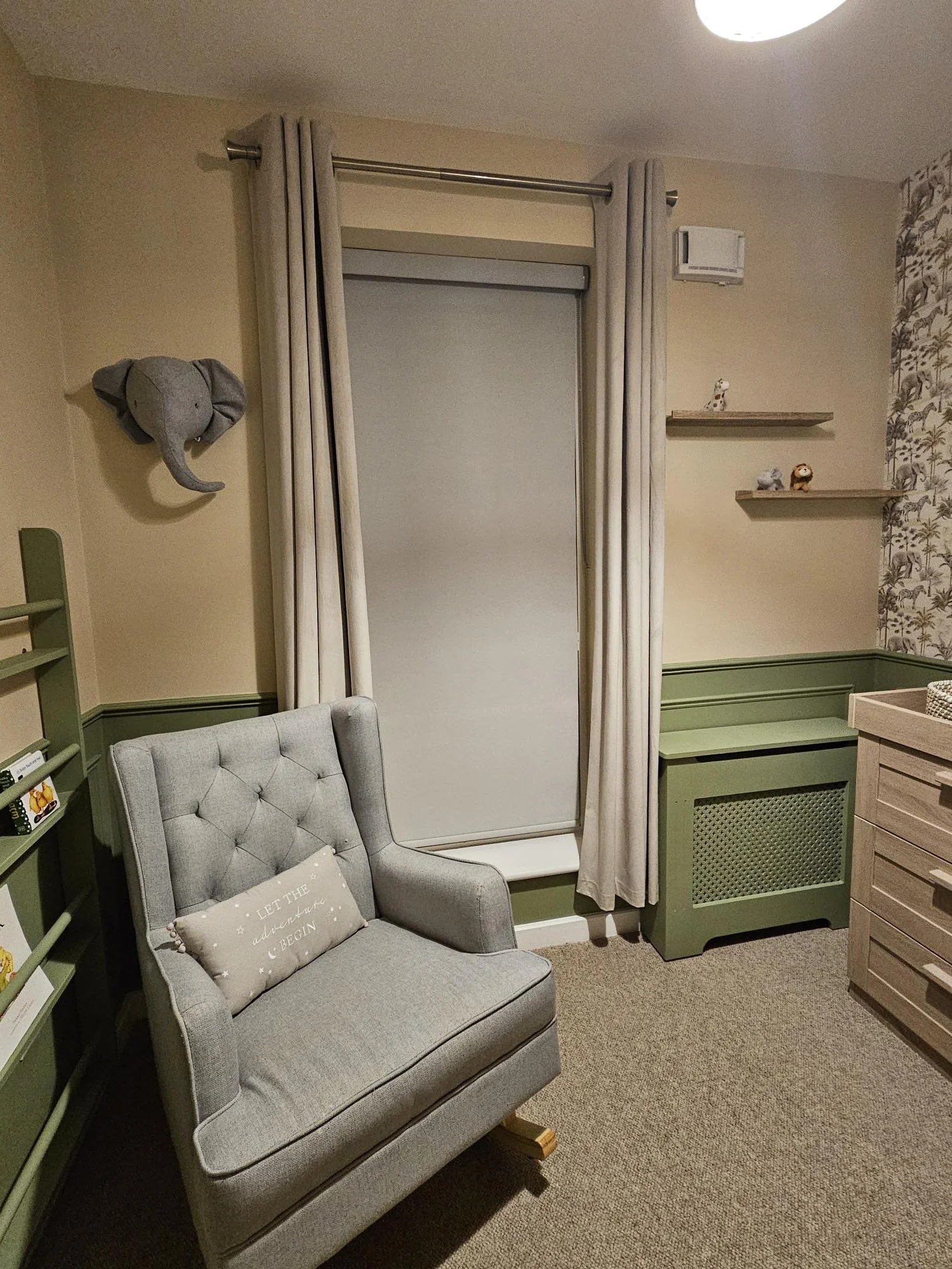 A cozy nursery with a light gray armchair with a beige pillow that says 'Let the adventure begin,' a small green wall-mounted storage unit, beige curtains, wall shelves with small plush toys, and a wall-mounted elephant head decoration by KF Designs.