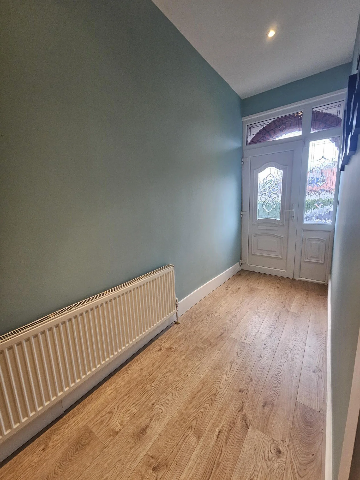 A small hallway with light blue walls, wooden flooring, a white radiator on the left wall, and a white front door with decorative glass panels, partially visible window on the right side, and a small ceiling light.