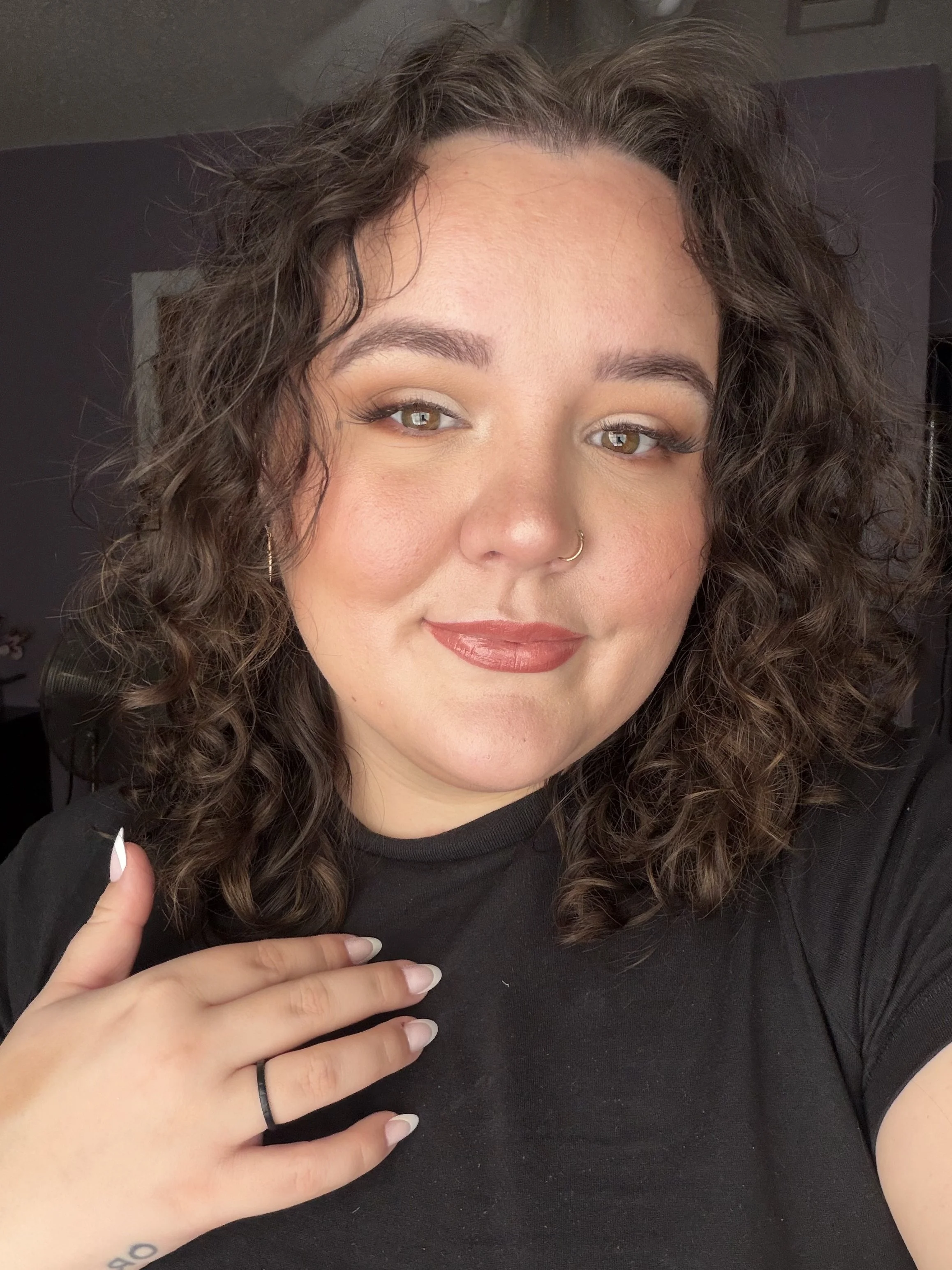 Close-up of a woman with curly hair, makeup, and a nose ring, holding her hand with a tattoo and a black hair tie, while pointing to her chest. She is wearing a black top.