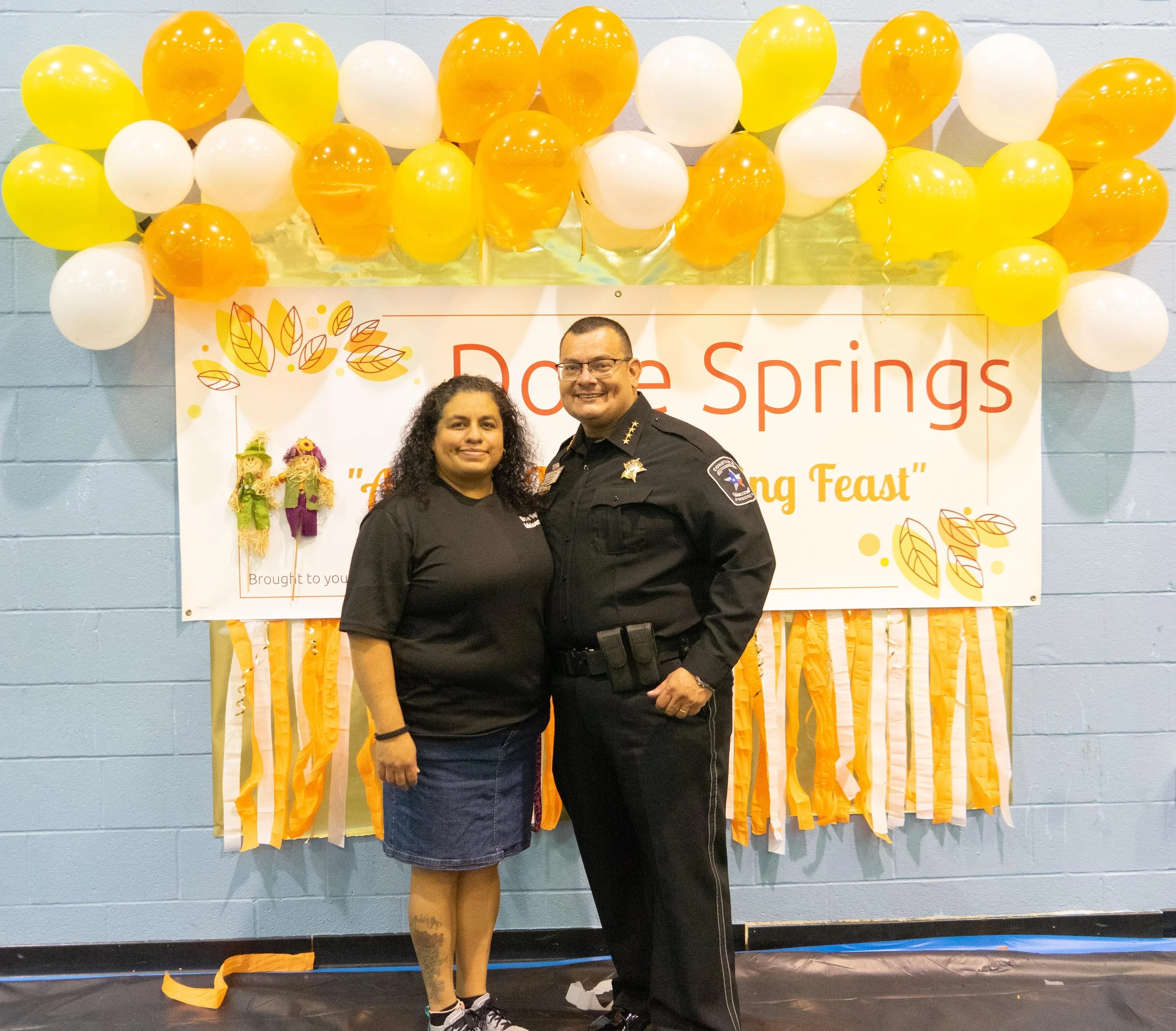 A woman and a police officer standing together in front of a decorated backdrop with yellow and white balloons, a banner reading 'Doe Springs, Fall Fest' and orange and yellow streamers.
