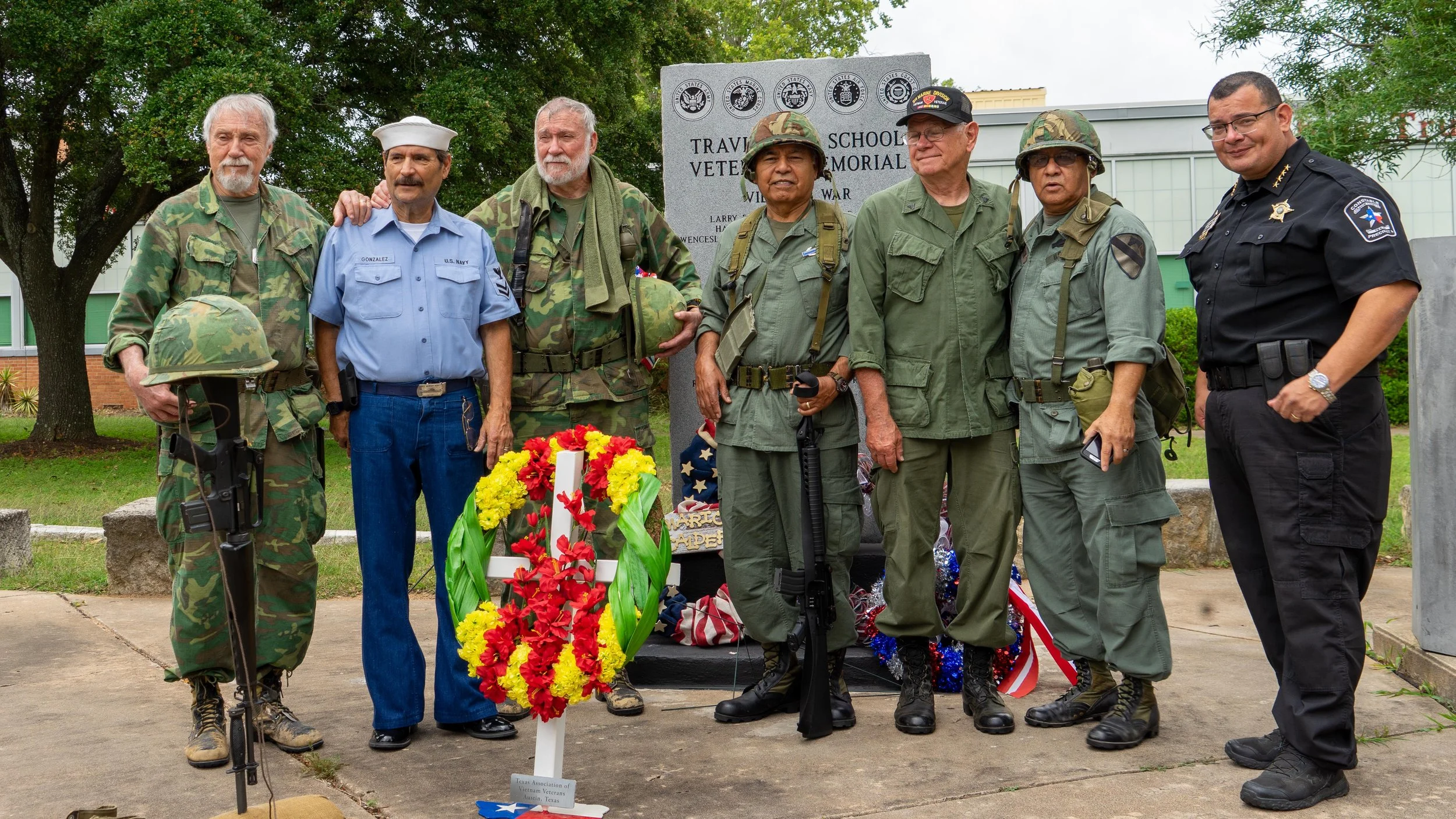 Group of veterans in military uniforms and a police officer standing at a memorial with a wreath of red, yellow, and green flowers, in front of a plaque honoring veterans, under a tree on a sidewalk.