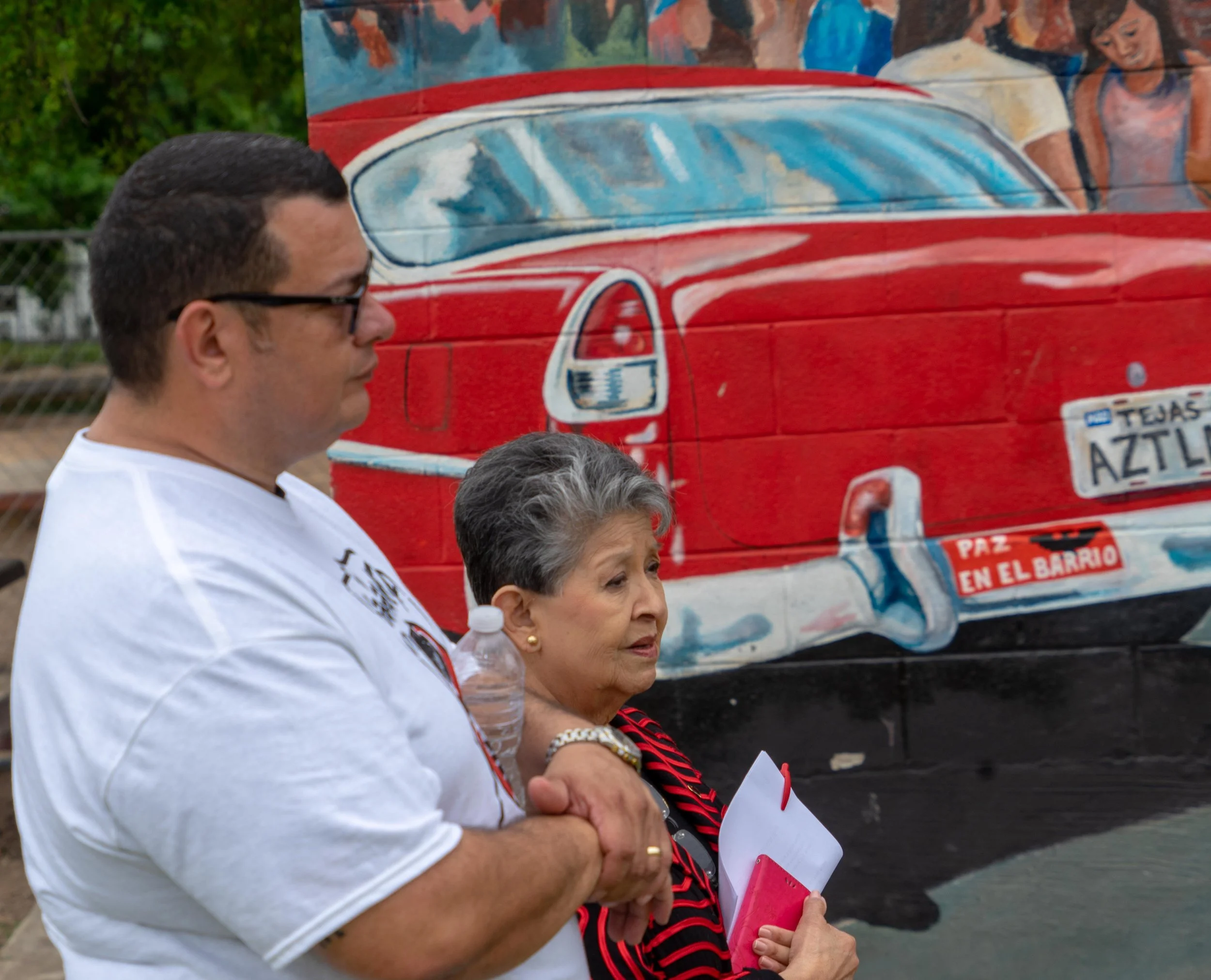A man and an elderly woman with gray hair are standing outdoors in front of a mural of a red vintage car painted on a brick wall. The woman is holding a notebook and a pen.