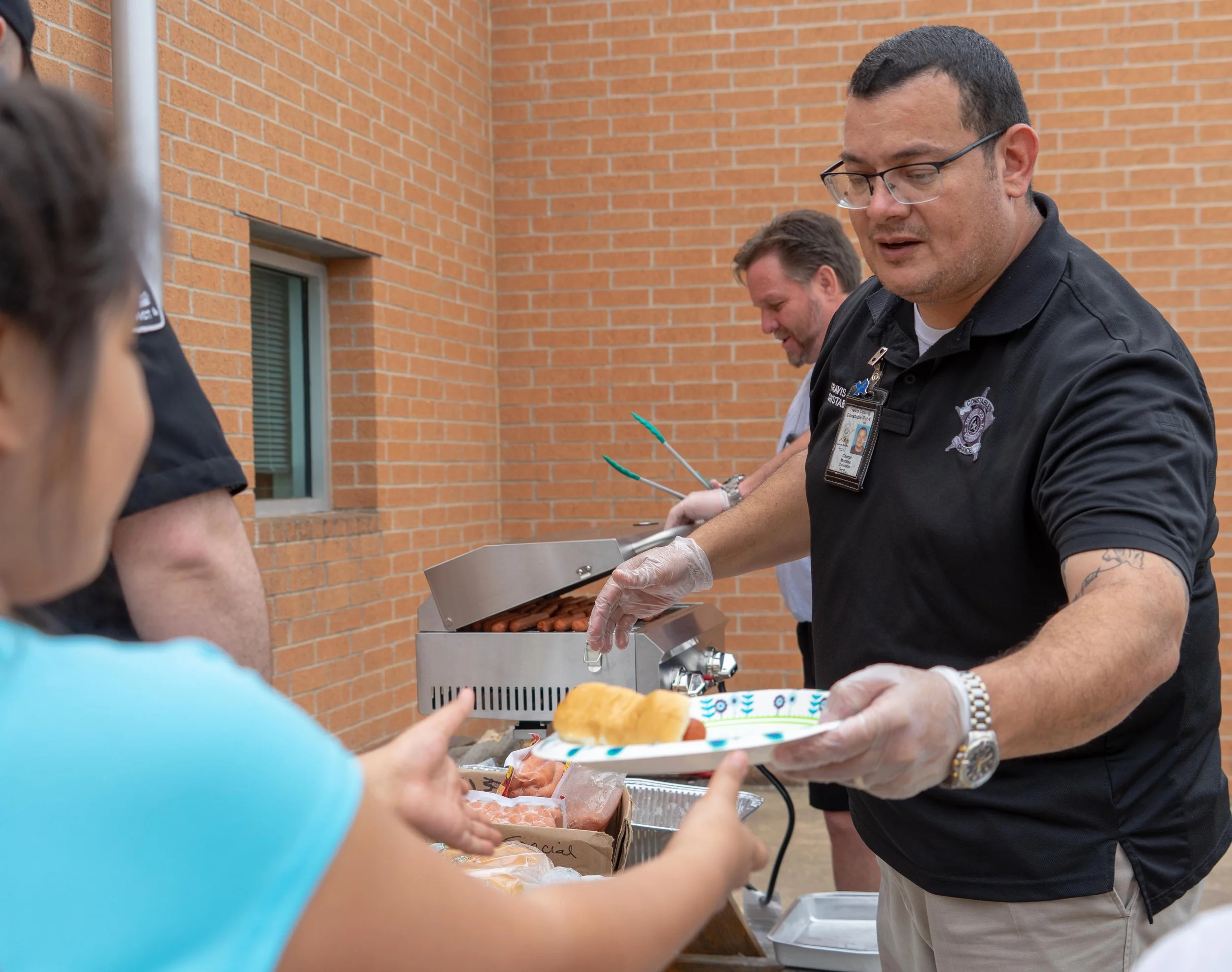 A man in a black shirt and glasses serving food from a grill to a child during a community event, with other people in the background.