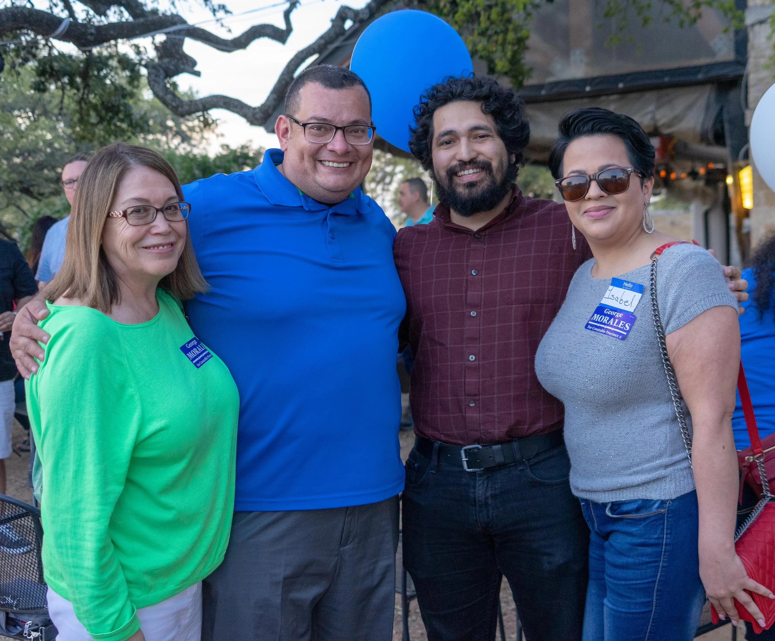 Four people standing together outdoors at a gathering, two women and two men, smiling at the camera, with balloons in the background.