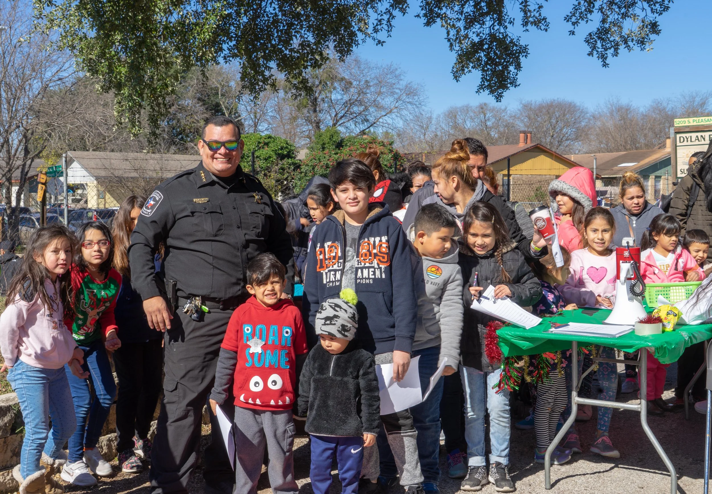 A group of children and adults gathered outdoors during daytime, with a police officer smiling at the camera, some children holding papers, and a table decorated for a festive event.