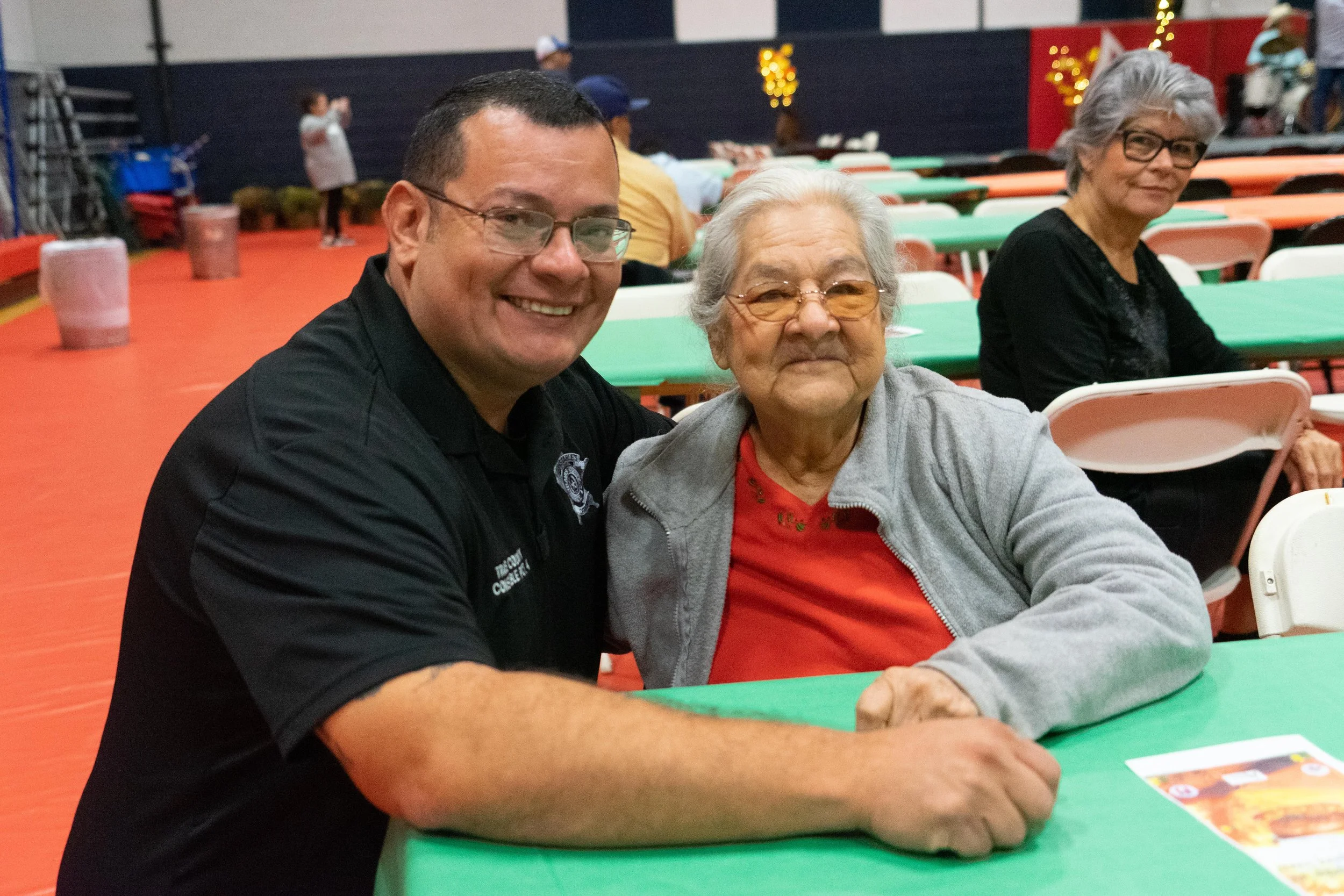 A man and an elderly woman sitting at a table in a decorated event hall, smiling at the camera while another woman sits in the background.