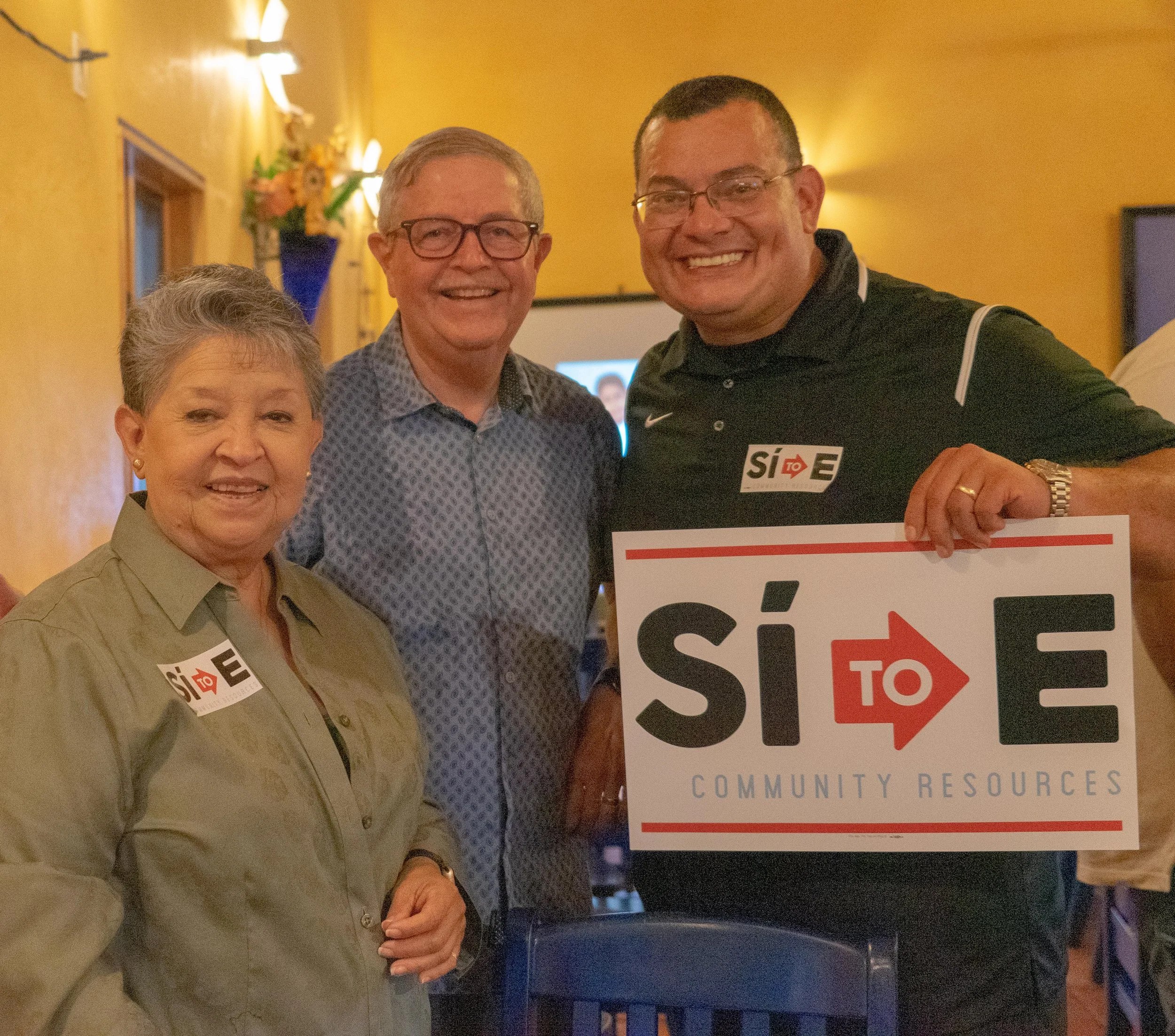 Three people standing together indoors, smiling at the camera. The man on the right is holding a sign that reads 'Sí to E' and 'Community Resources.' The woman on the left is wearing a beige shirt with a name tag, and the man in the middle is wearing