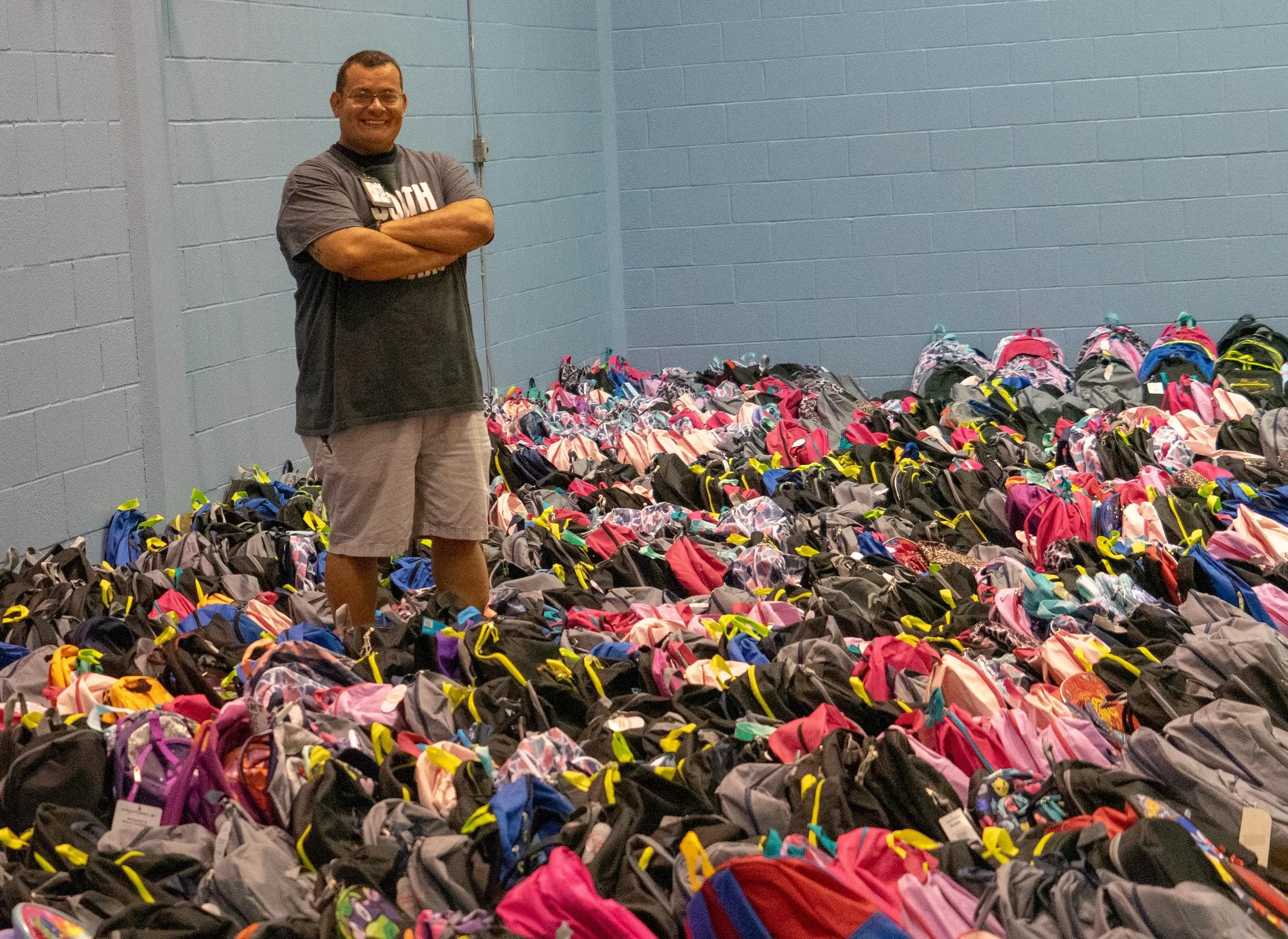 A man standing with crossed arms beside numerous backpacks organized on the floor, in a room with blue walls.