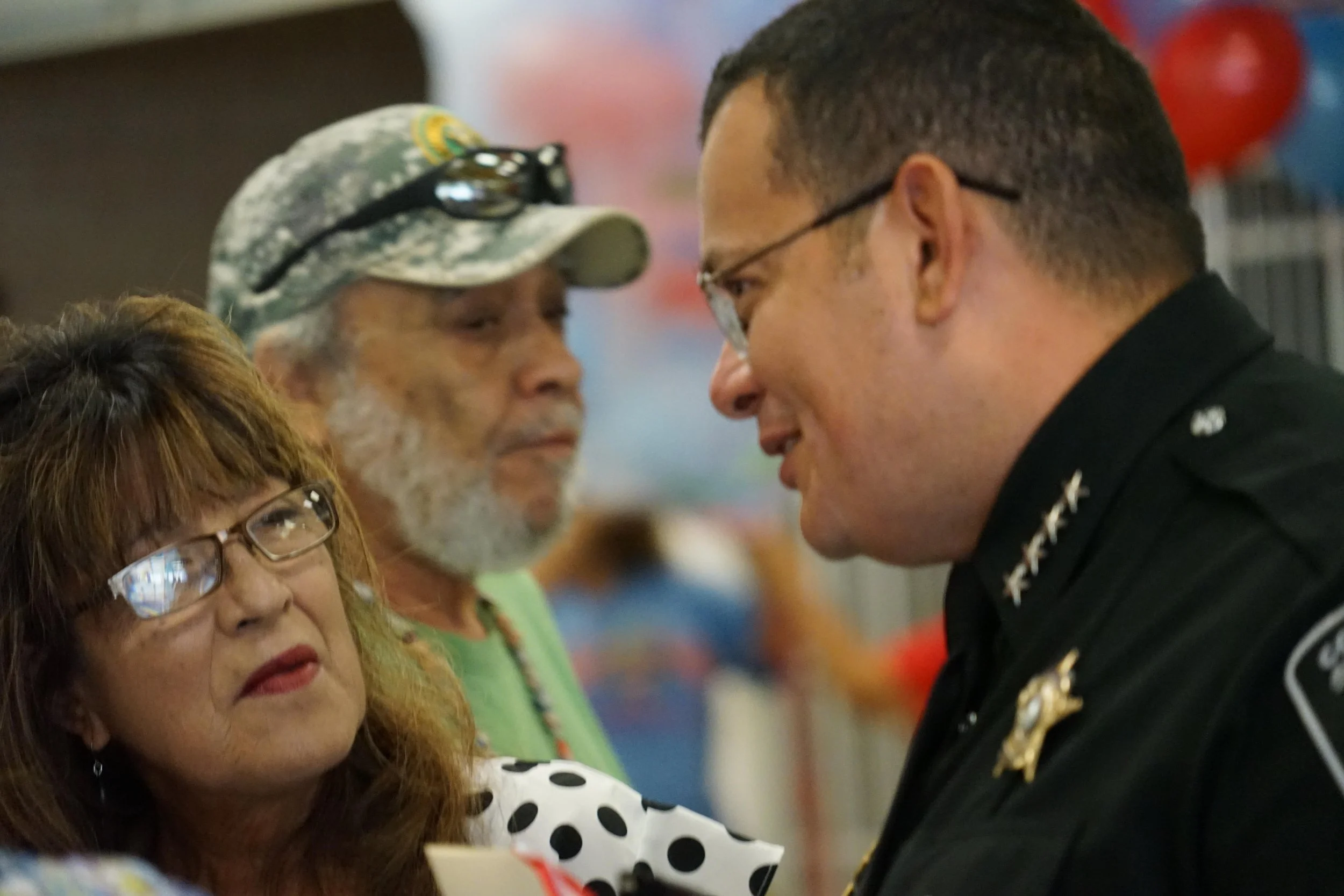 A woman with glasses and red hair looking at a man in a police uniform, who is smiling and leaning in to speak with her. An older woman with a beard and glasses, wearing a camouflage hat, is in the background.