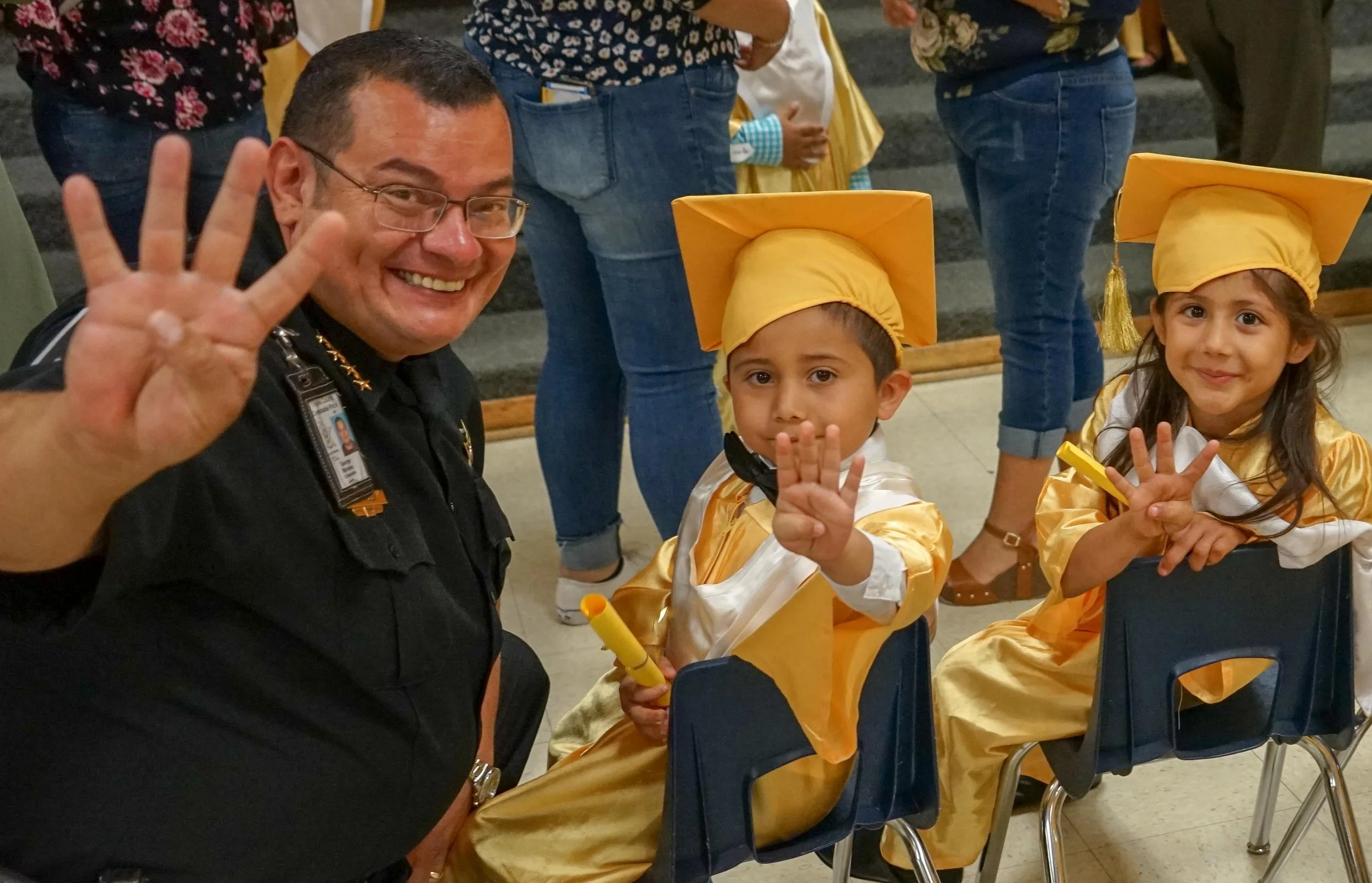 A smiling man in uniform with glasses is waving at the camera. Two young children dressed in gold graduation caps and gowns are sitting on chairs, waving and smiling at the camera. The boy holds a yellow diploma. The girl is holding a yellow item, po