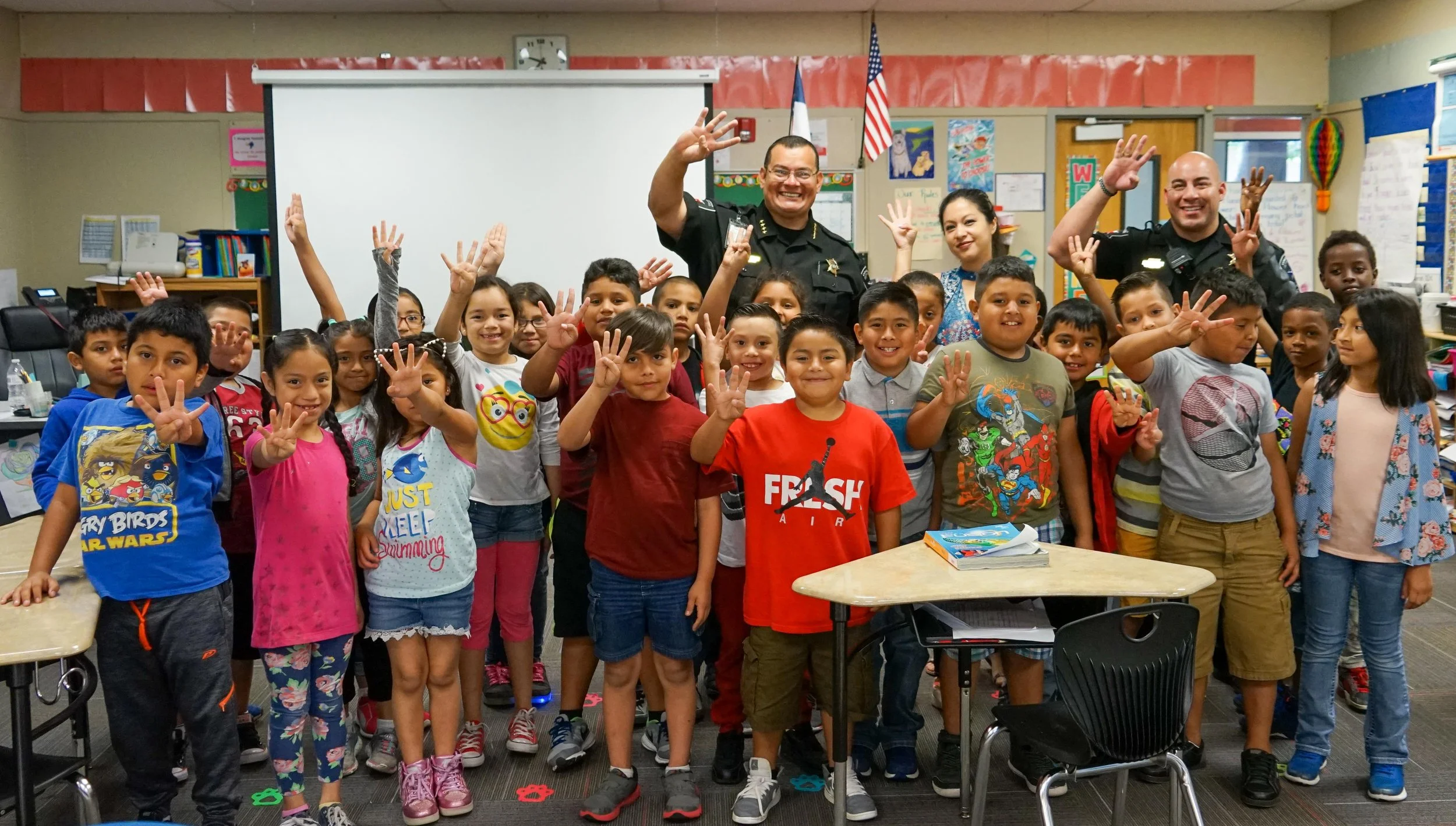 A group of elementary school children and police officers indoors, smiling and waving at the camera.