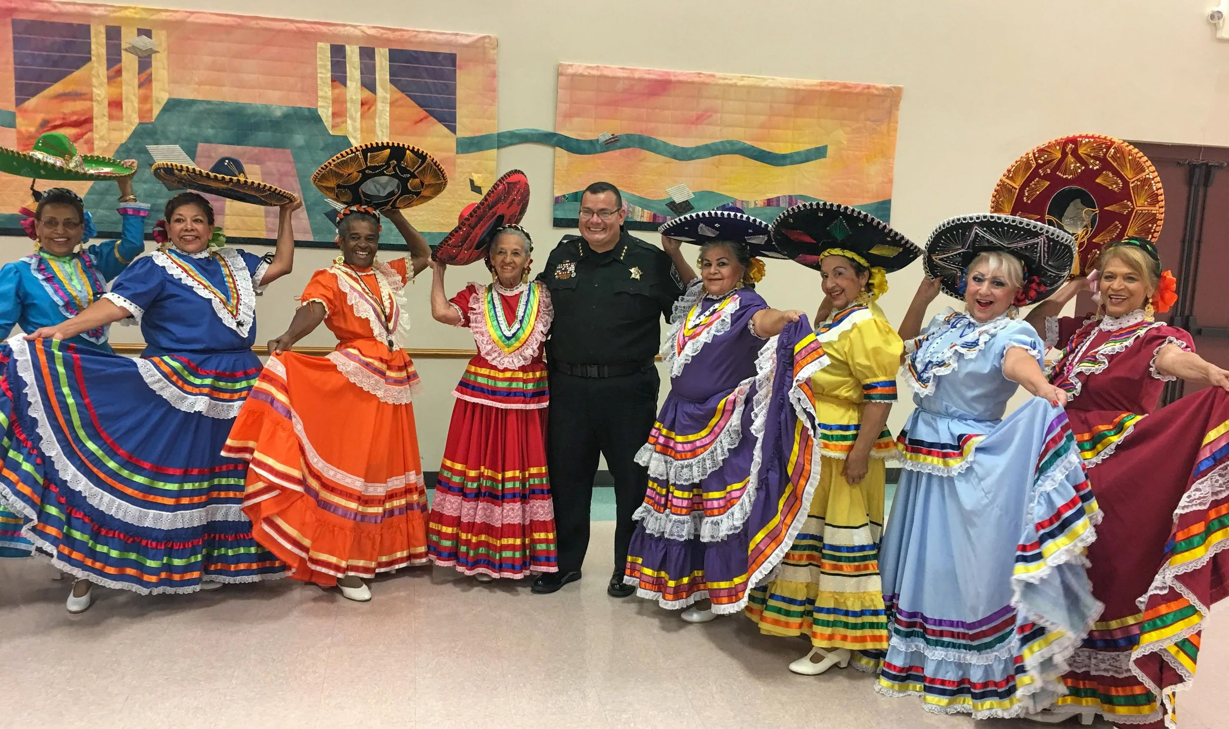 Group of women dressed in traditional colorful Mexican dresses holding sombreros, standing with a police officer indoors in front of abstract wall art.