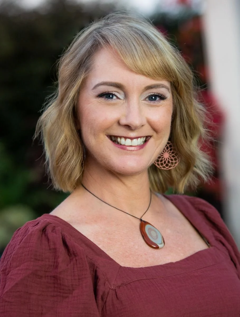 A smiling woman with shoulder-length blonde hair wearing a maroon top, a colorful pendant necklace, and pink earrings, standing outdoors with a blurred background.