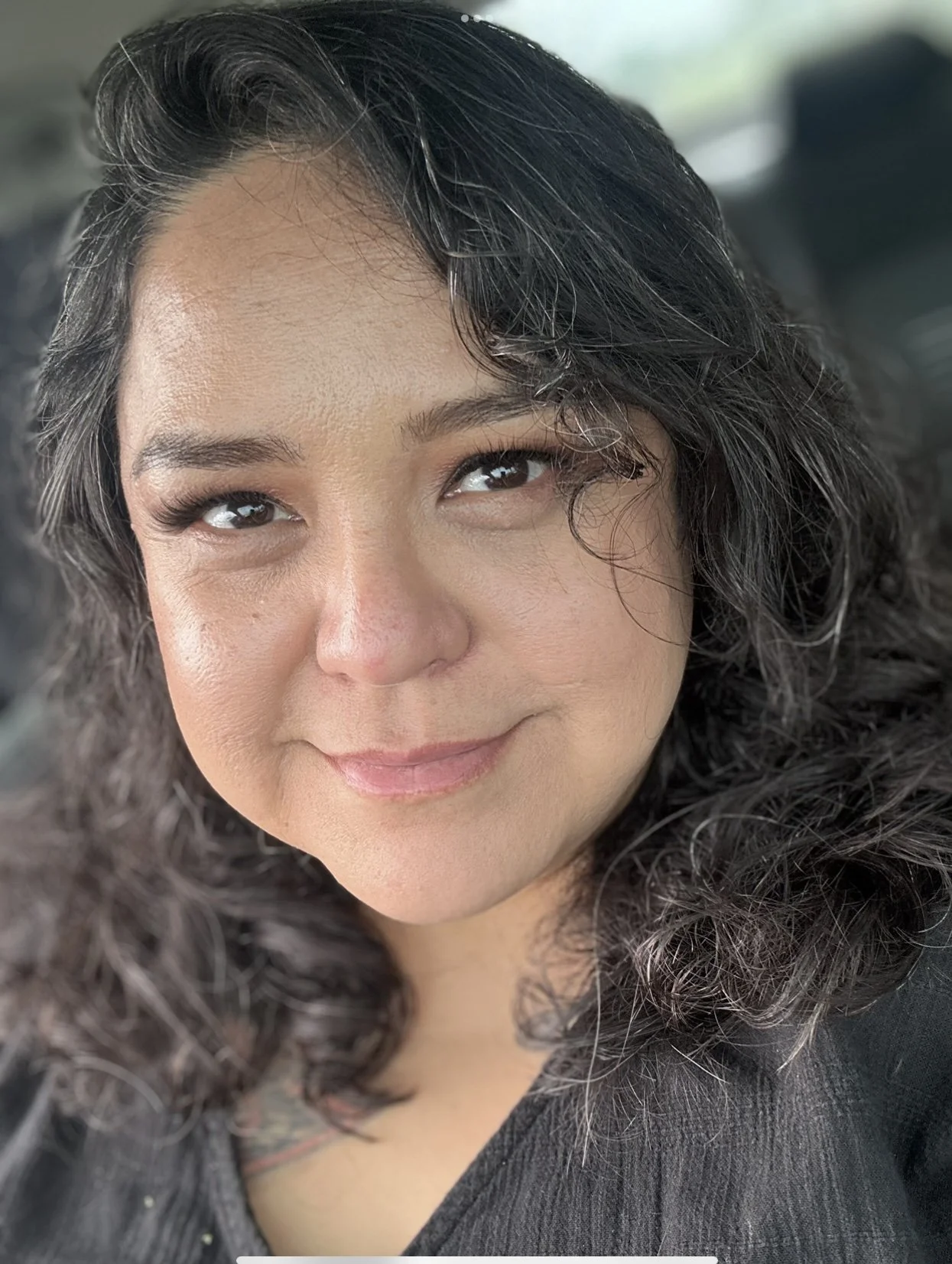 A woman with medium skin tone and curly black hair taking a selfie, smiling softly and looking slightly to the side.